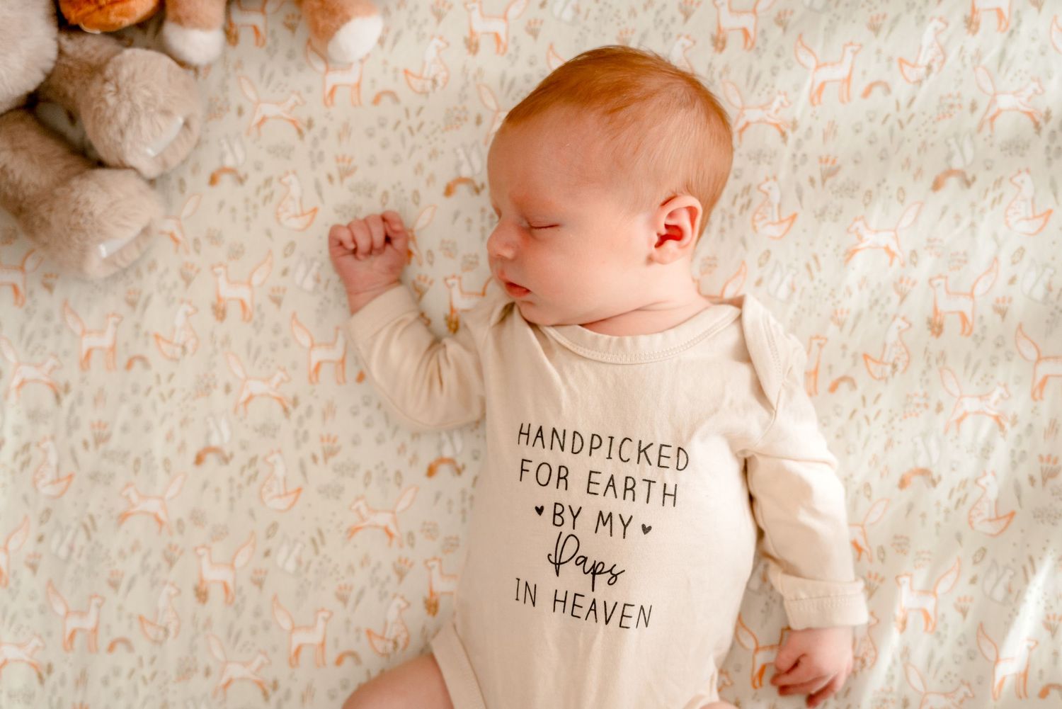 Sleeping newborn baby in cream onesie reading Handpicked For Earth By My Pops In Heaven on floral blanket.