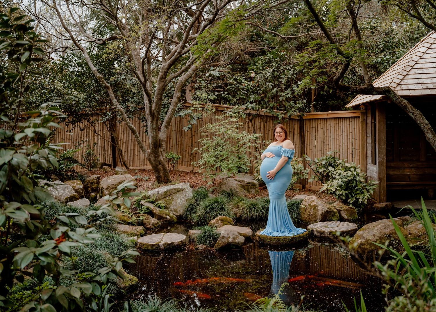Pregnant woman in blue gown standing on stones over koi pond in lush garden for maternity photo.