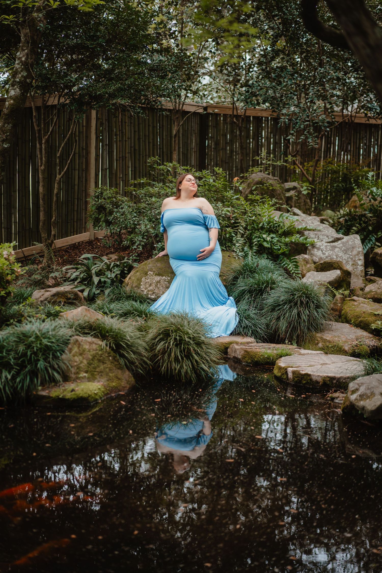 A pregnant woman in a blue dress stands in a garden with a wooden fence and lush vegetation, reflected in a pond.