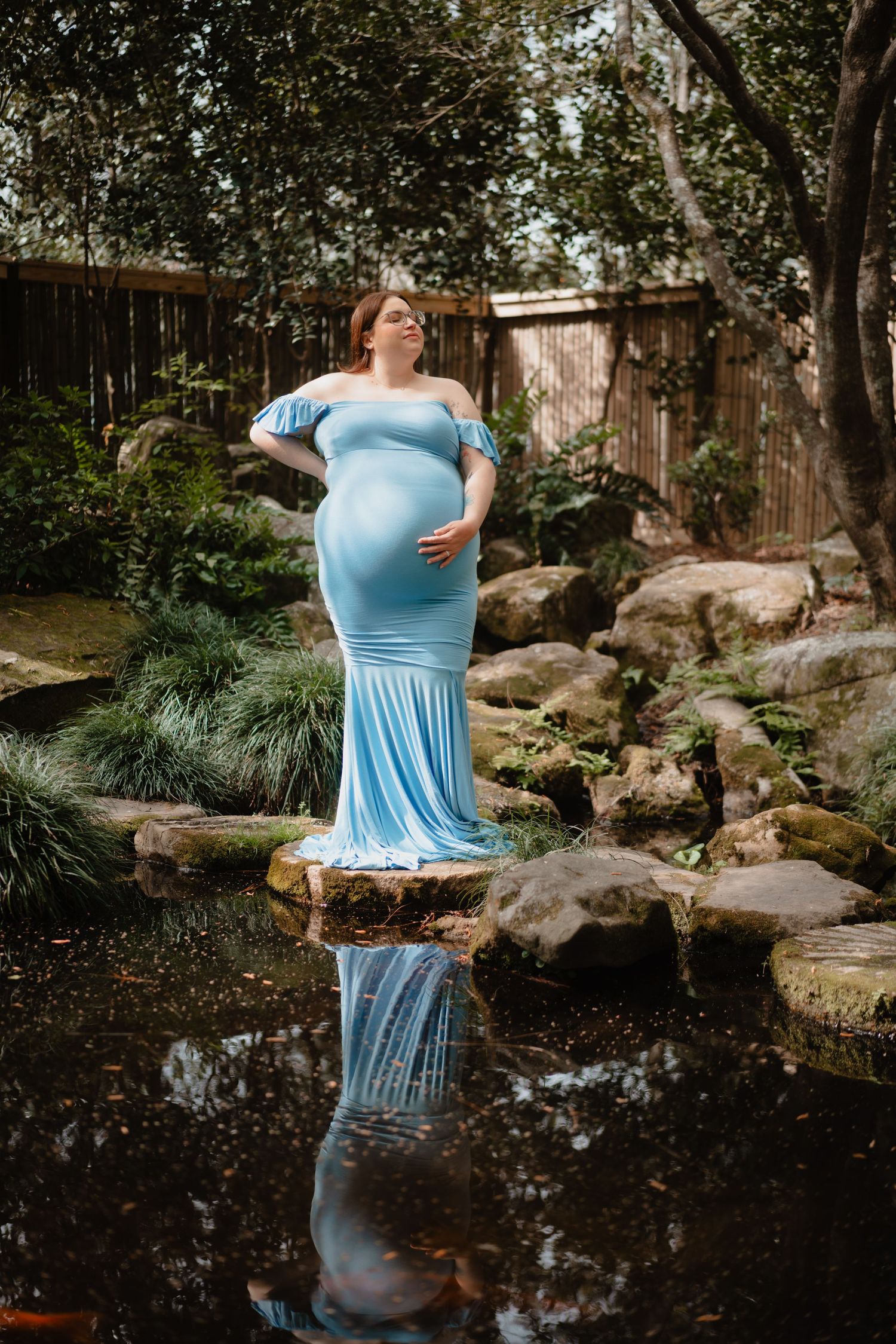 A pregnant woman in a blue dress stands in a lush garden, her reflection visible in the water below.