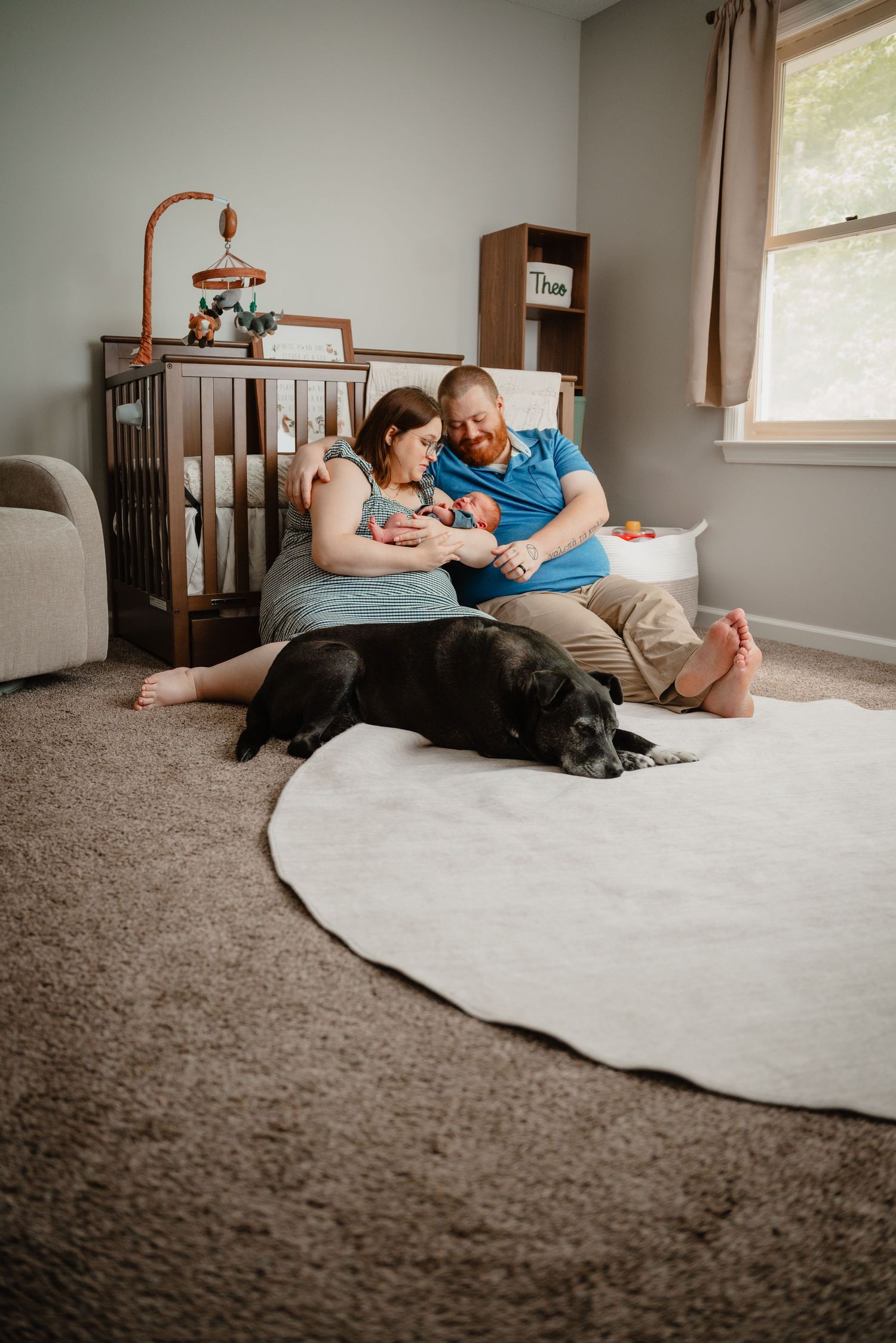 A couple sits on nursery floor with newborn baby and black Labrador dog, with crib and mobile in background.
