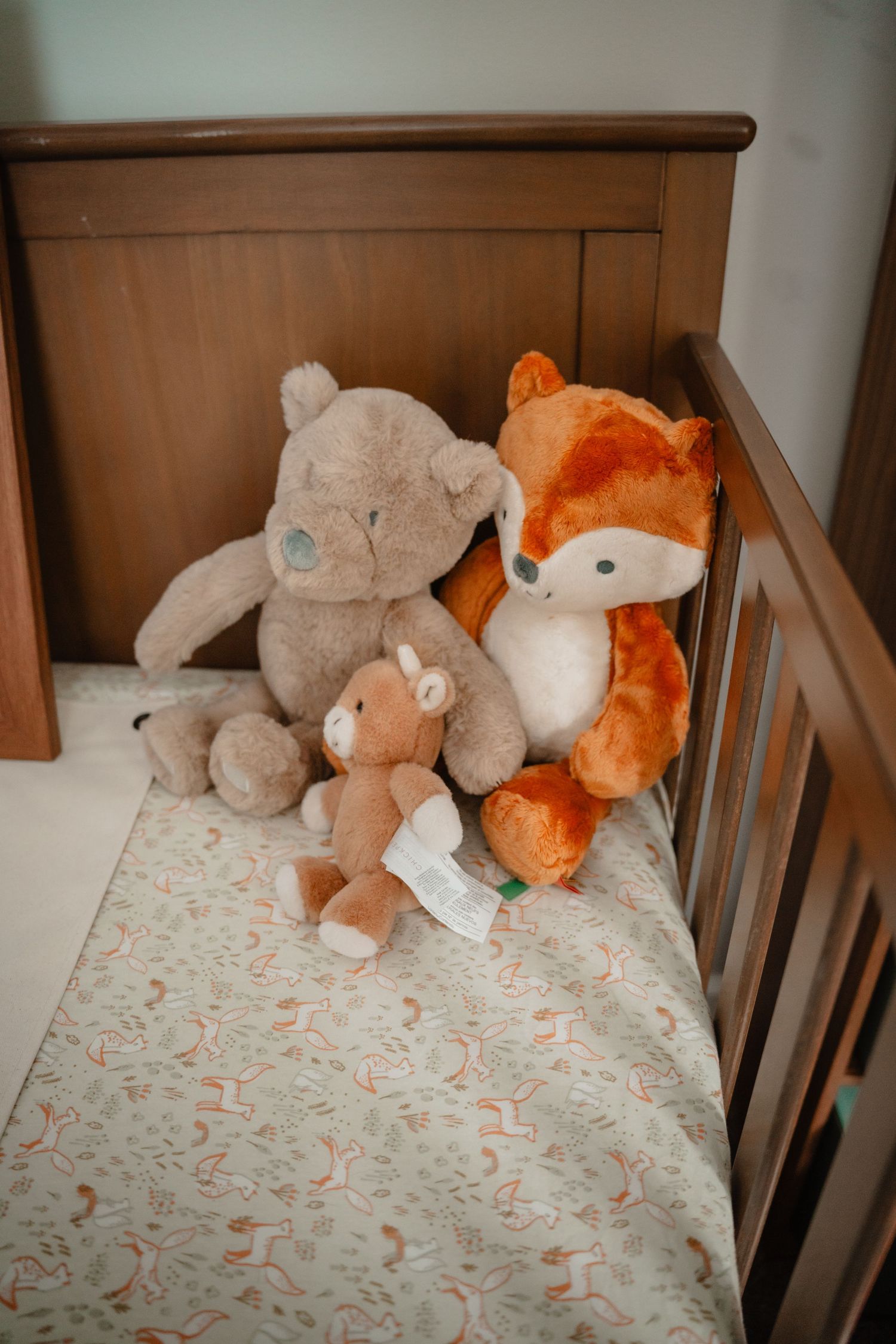 Nursery decor of three stuffed animals, two teddy bears and an orange fox plush, sitting in a wooden baby crib with a patterned sheet.