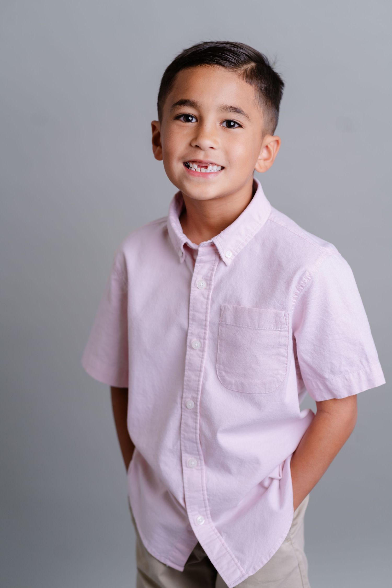 A young child poses playfully in a light pink button-down shirt against a gray background.