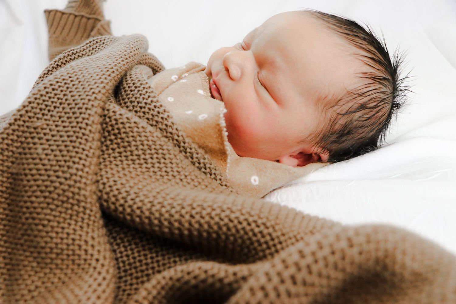 A sleeping newborn baby wrapped in a cozy brown knit blanket rests peacefully on white bedding.