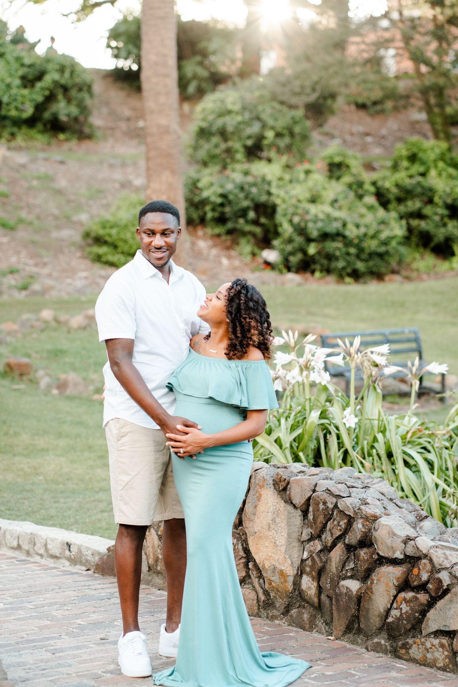 A couple embraces in a garden setting with stone wall and palm trees, wearing white shirt and mint green formal dress.