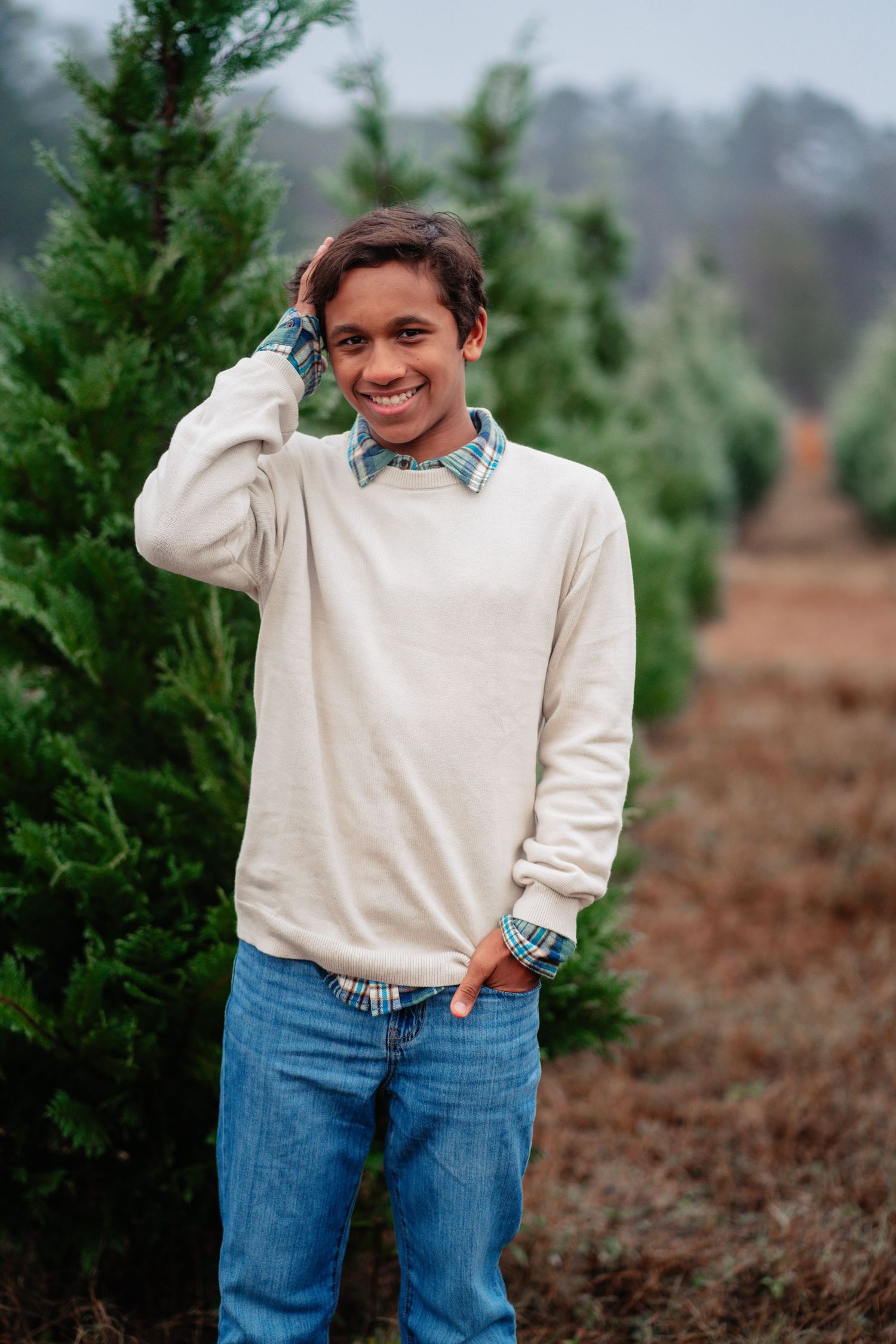 Person in cream sweater and jeans poses playfully against backdrop of evergreen trees at Christmas tree farm.