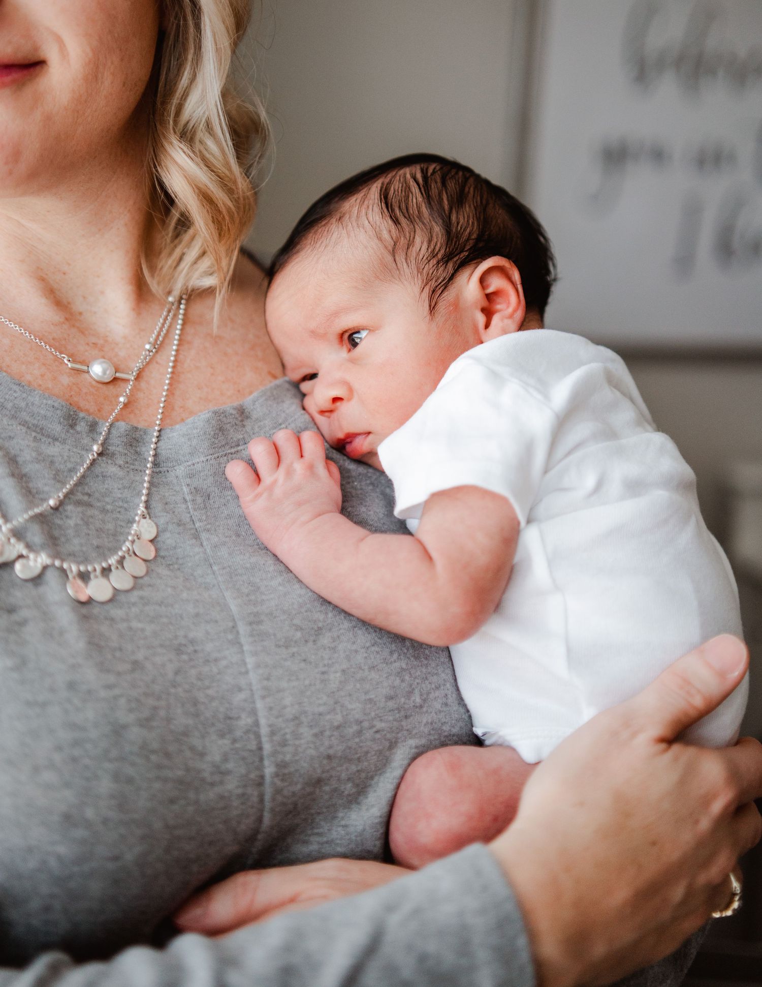 Newborn baby in white onesie resting against parent wearing gray shirt.