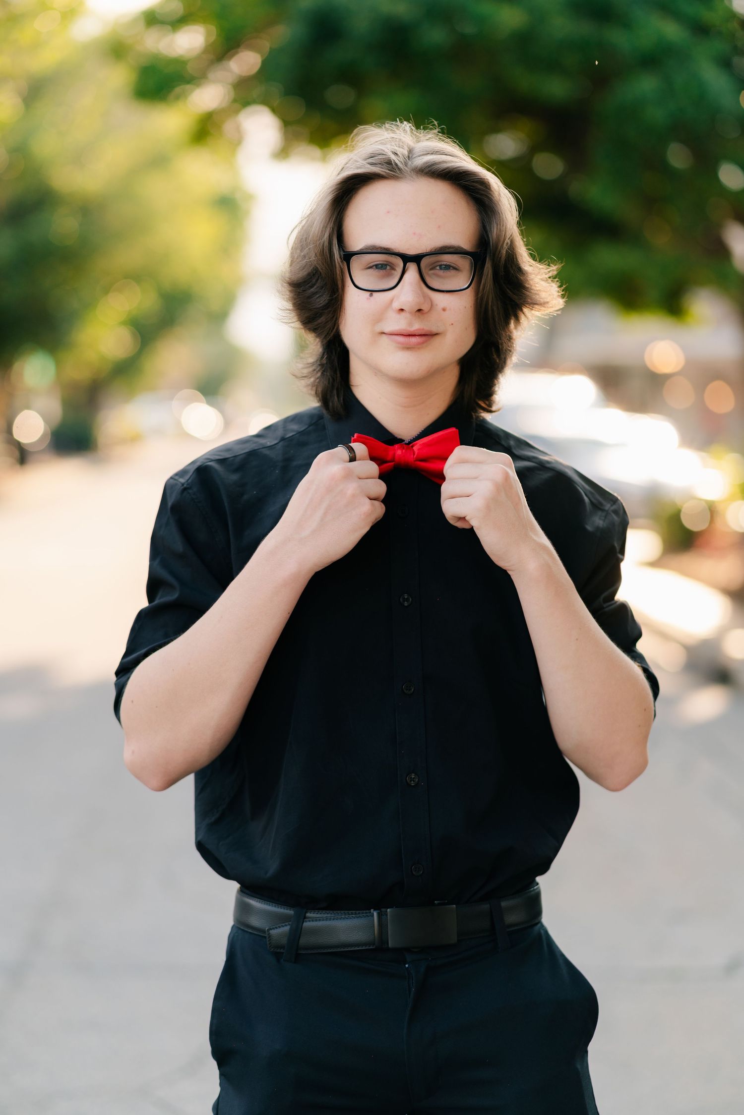 A person in a black shirt and red bowtie adjusts their tie while standing outdoors with a blurred background.