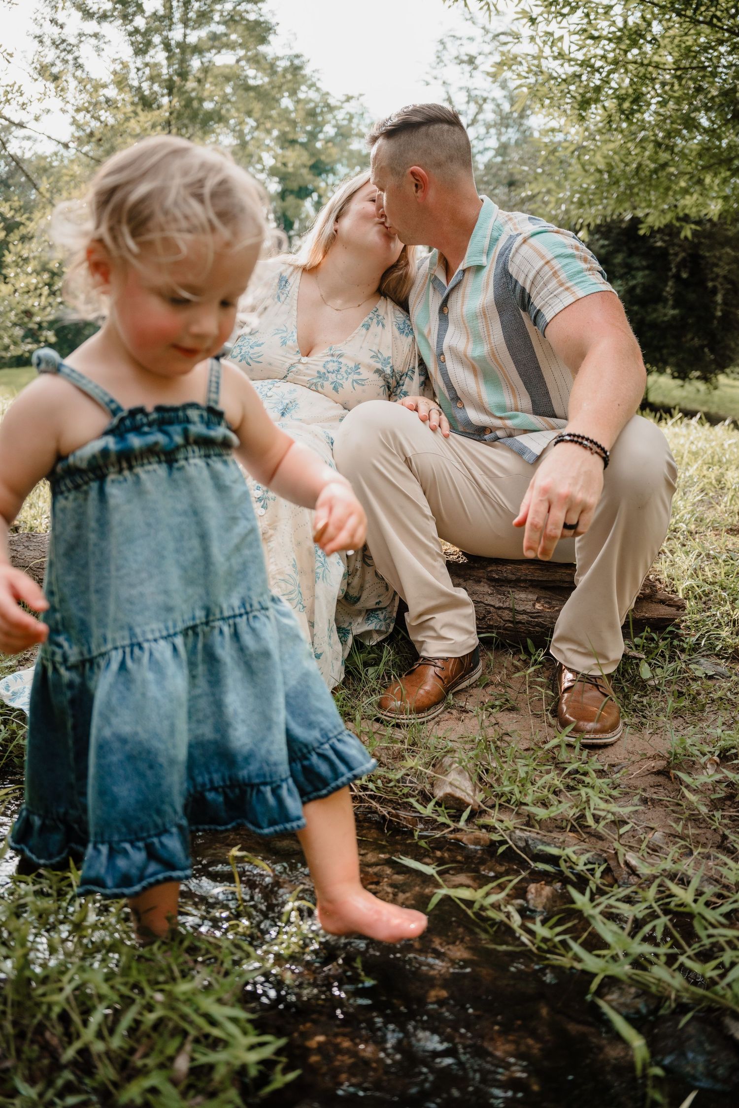 A young girl in a denim dress plays near a stream while her parents share a kiss on a log in a sunny garden.