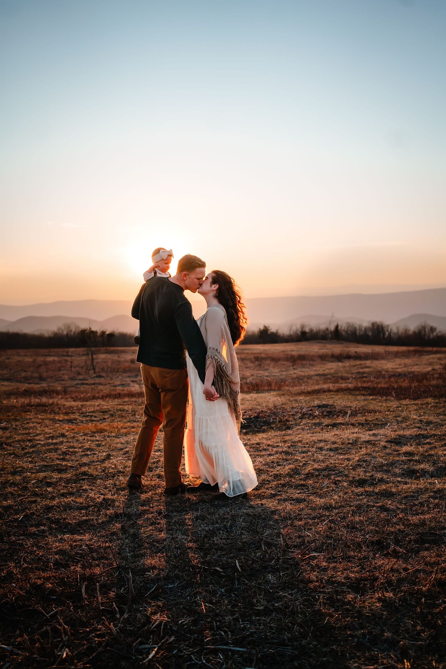 Couple sharing a romantic kiss at golden sunset in an open field with mountain views in the background.