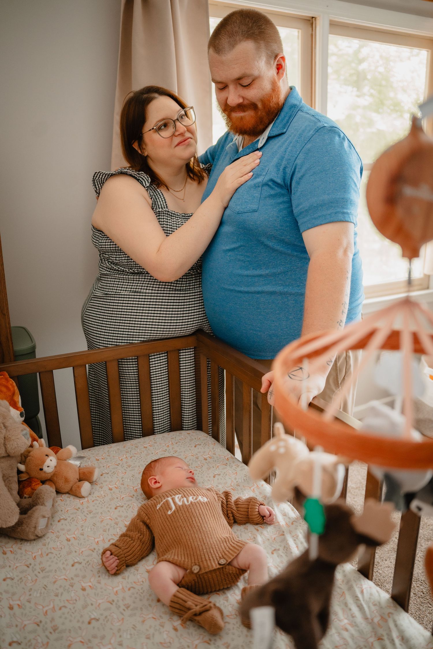 A couple smiles lovingly at their newborn baby lying in a wooden crib in a softly lit nursery room during in-home newborn session.