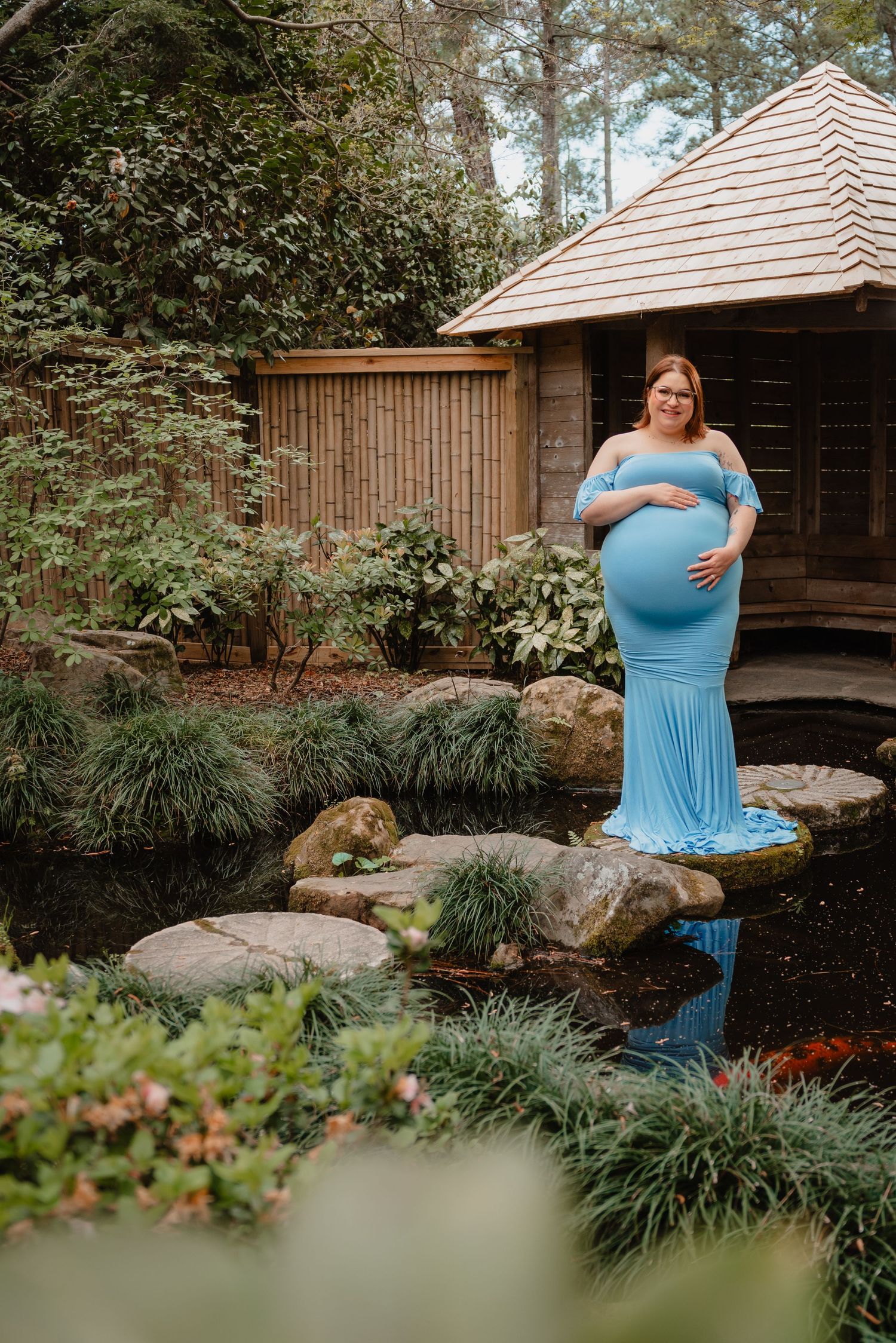 A pregnant woman in a blue dress stands in a lush garden with a wooden structure in the background.