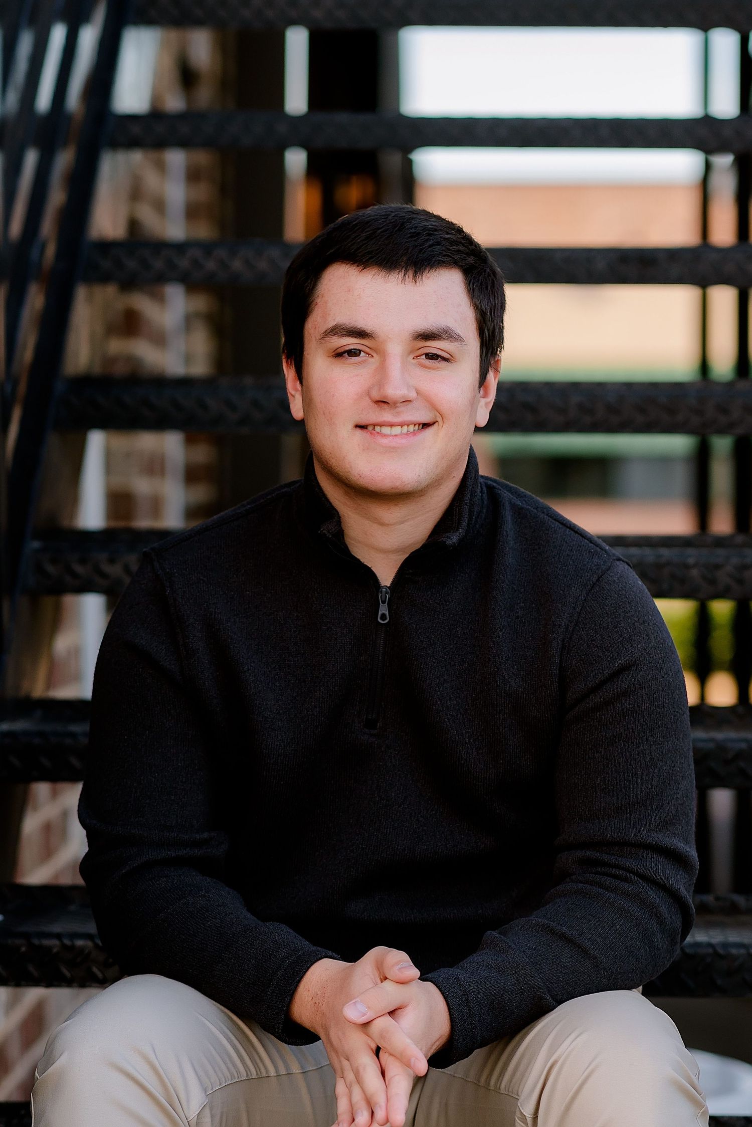 A person wearing a black sweater sits casually on steps with a warm smile in an outdoor setting.