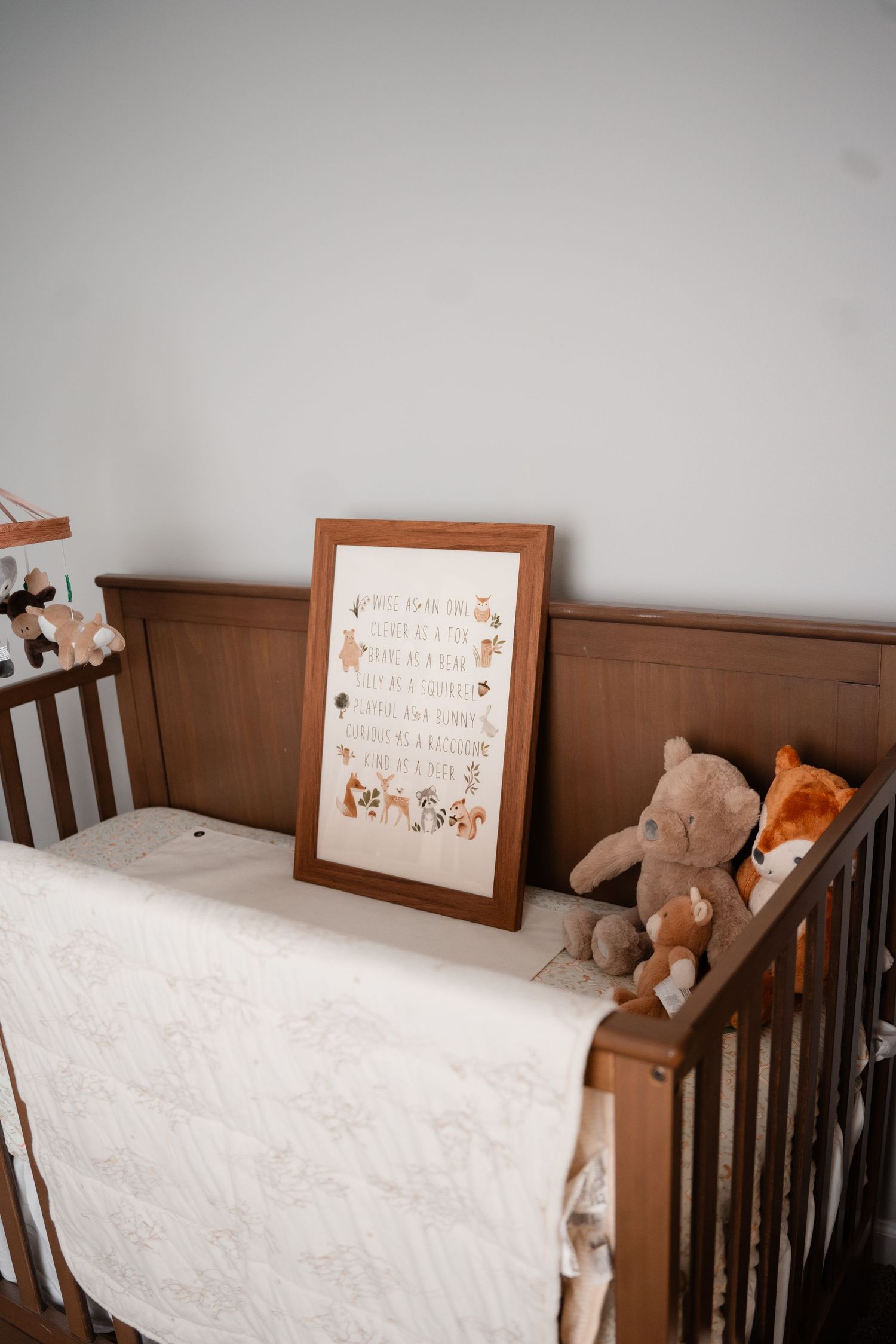 Wooden baby crib with white quilt, stuffed animal toys, and a framed woodland nursery print leaning against the side.
