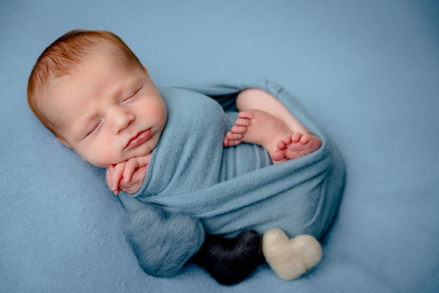Newborn baby sleeping peacefully wrapped in a blue swaddle blanket with two small heart-shaped toys nearby.