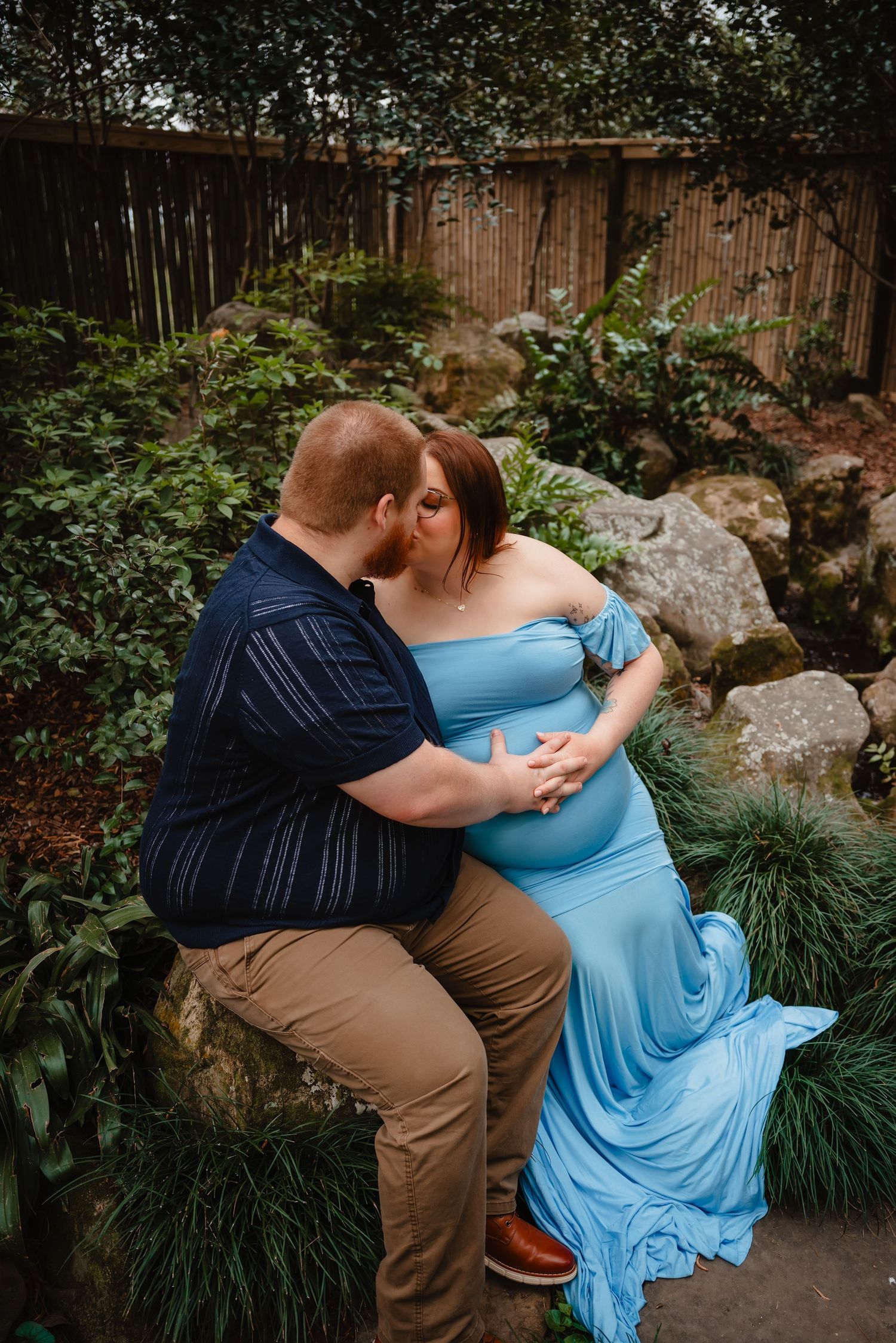 A pregnant woman in a blue dress is embraced by her partner in a lush, outdoor garden setting.