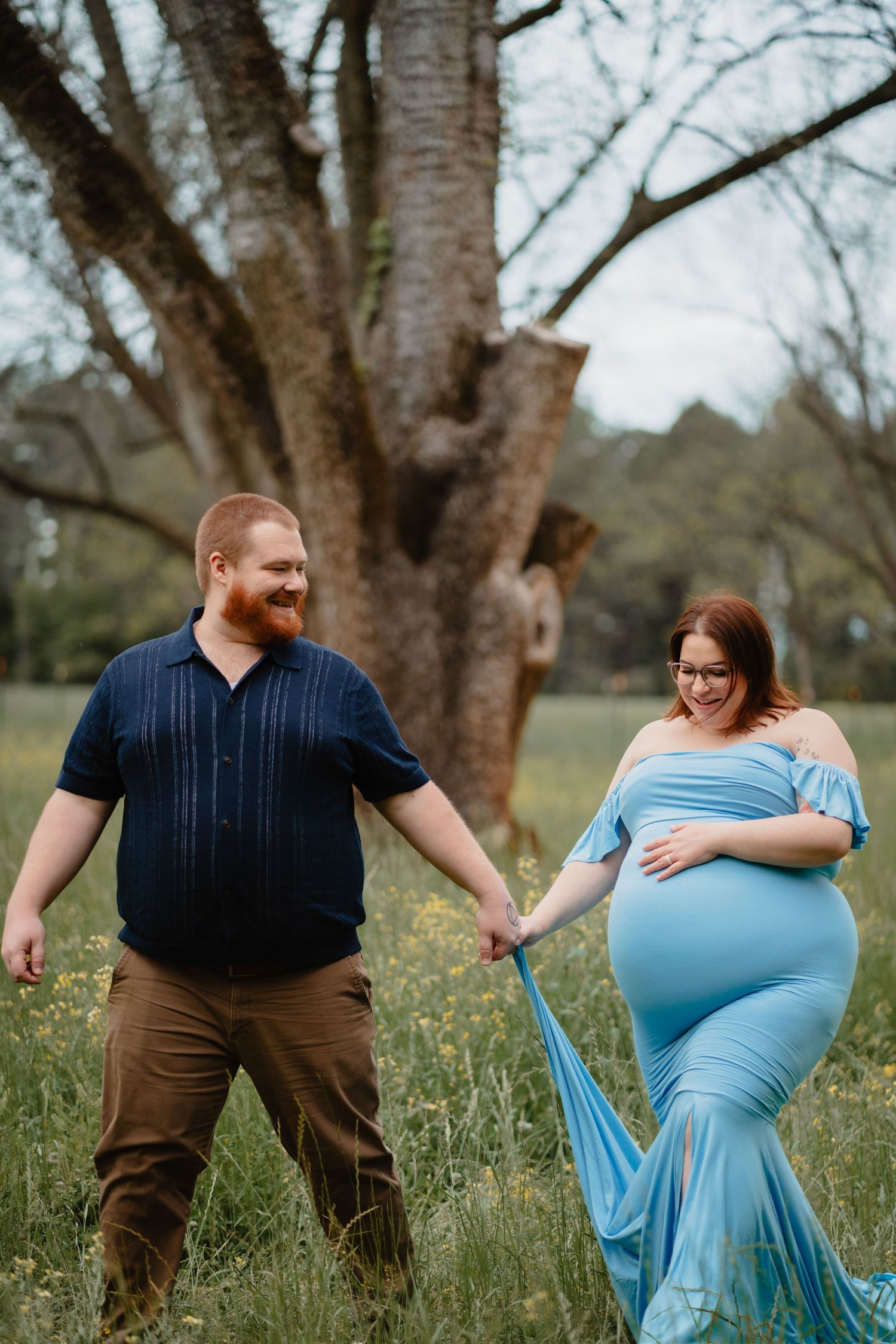 A pregnant woman in a blue dress stands with her husband in a grassy field surrounded by trees.