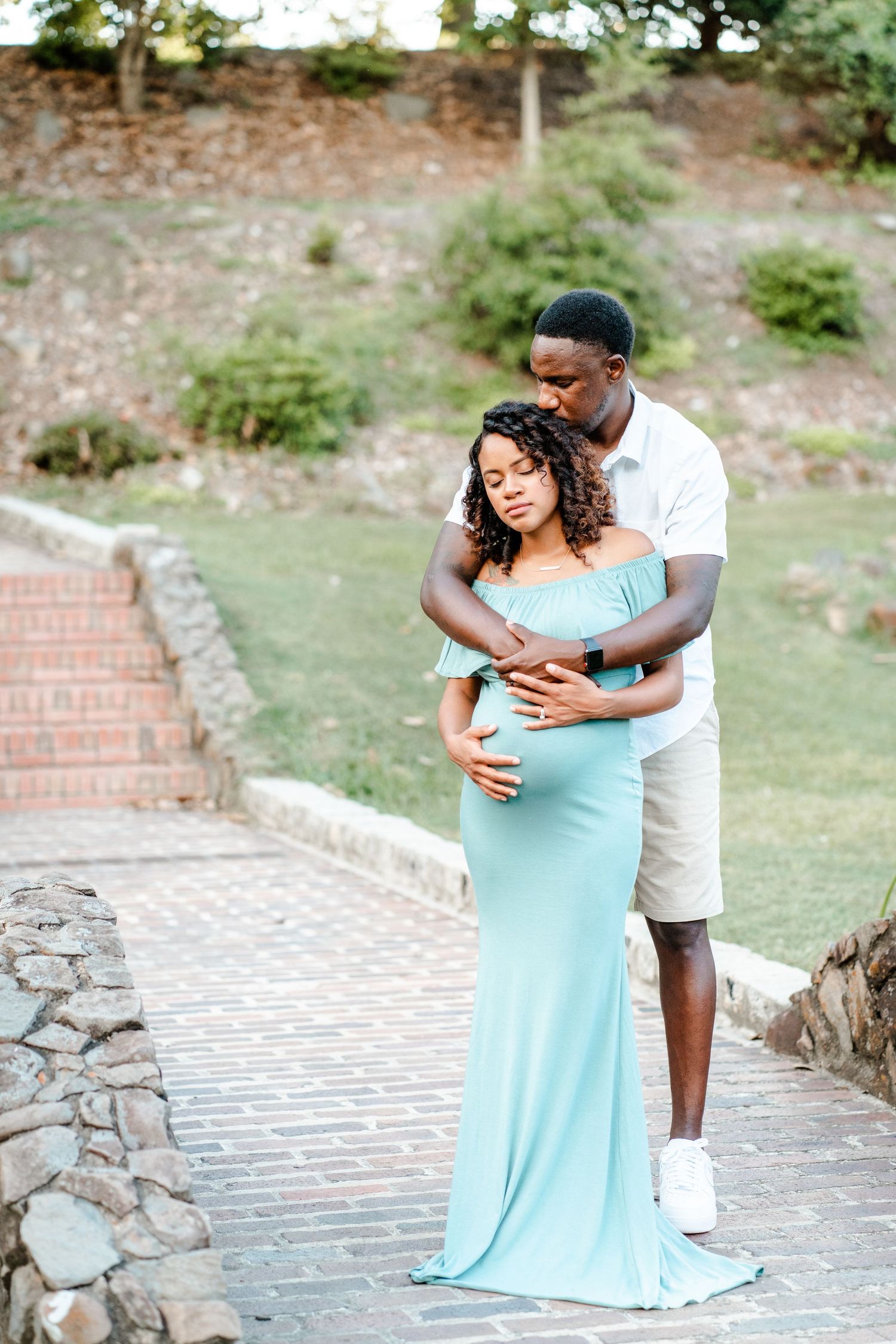 A romantic maternity photoshoot captured on a stone path with a couple embracing in matching white and blue outfits.