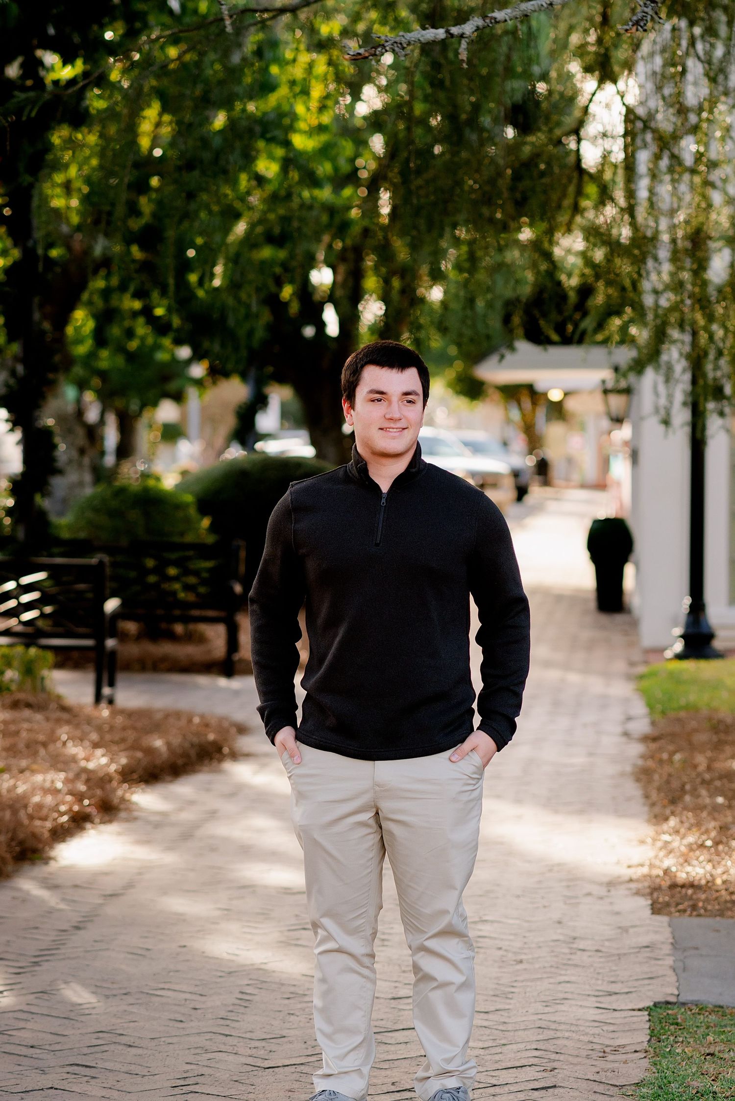 Person in black sweater and khaki pants standing on brick pathway surrounded by trees and greenery.