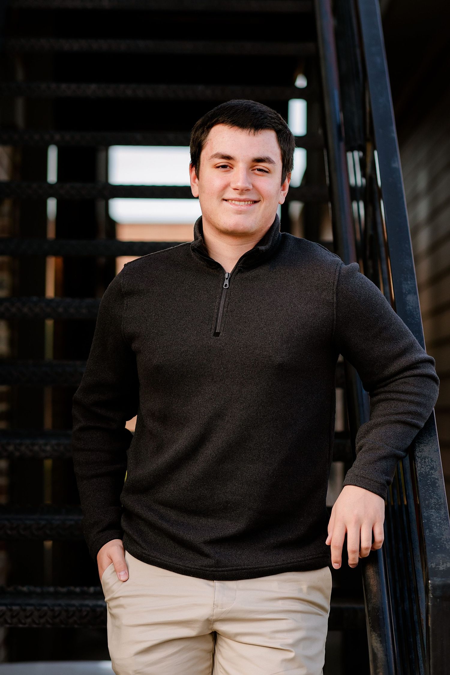 Person in black quarter-zip sweater and khaki pants poses against dark industrial background in multiple shots.