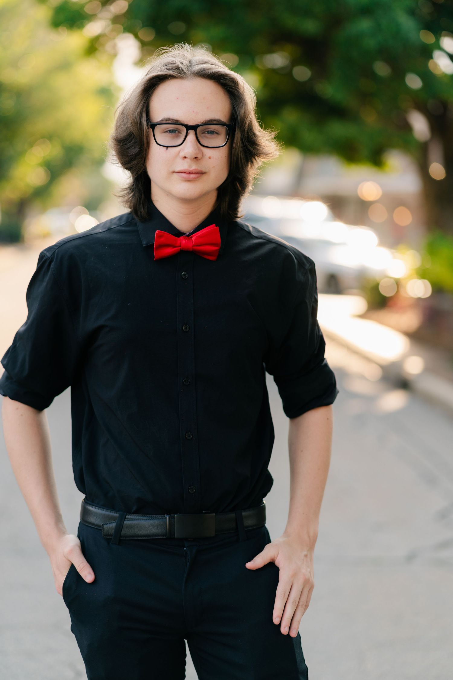 A figure in black attire and red bowtie poses confidently against a sunlit outdoor background.