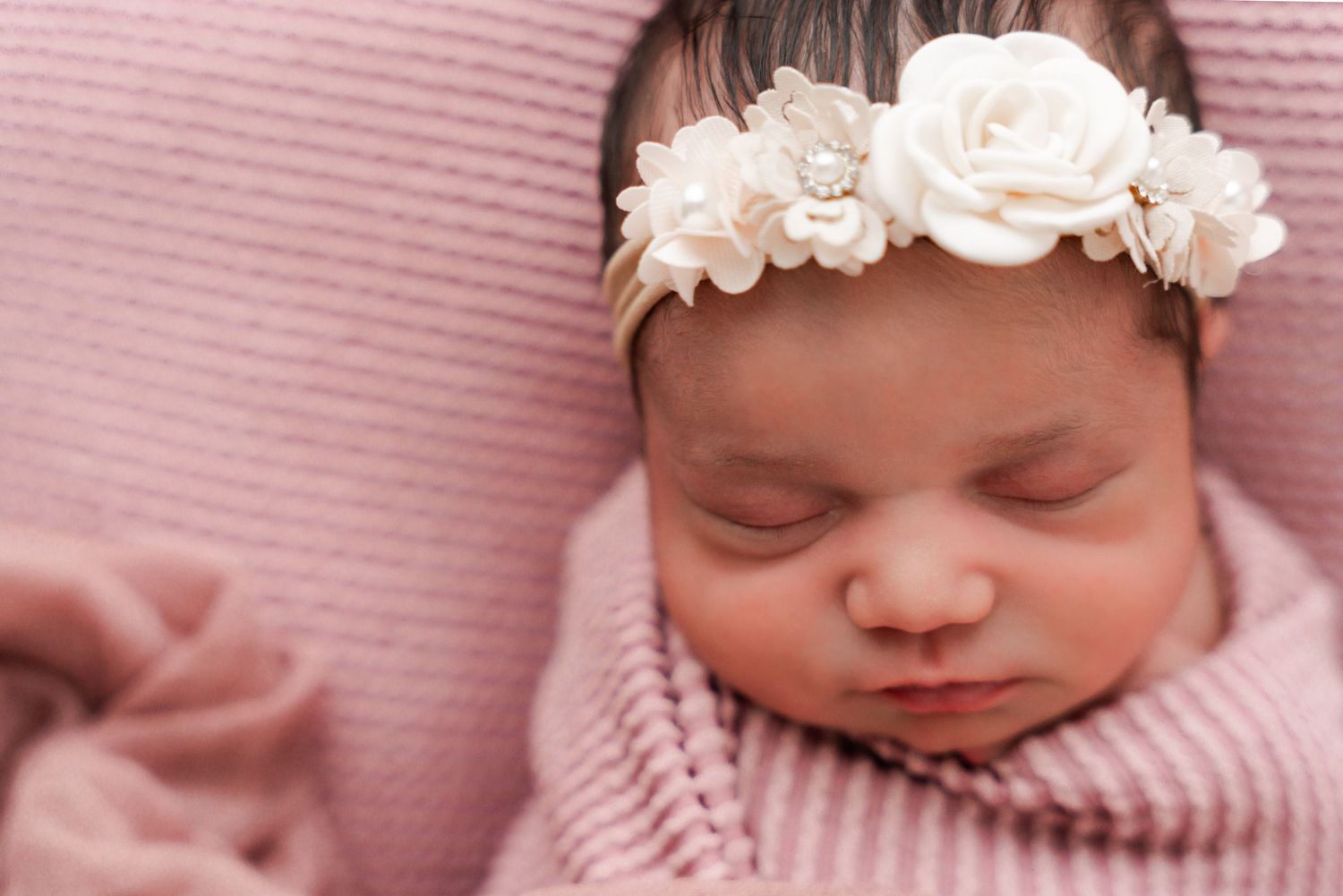 Sleeping newborn baby girl wrapped in pink with a white floral headband.