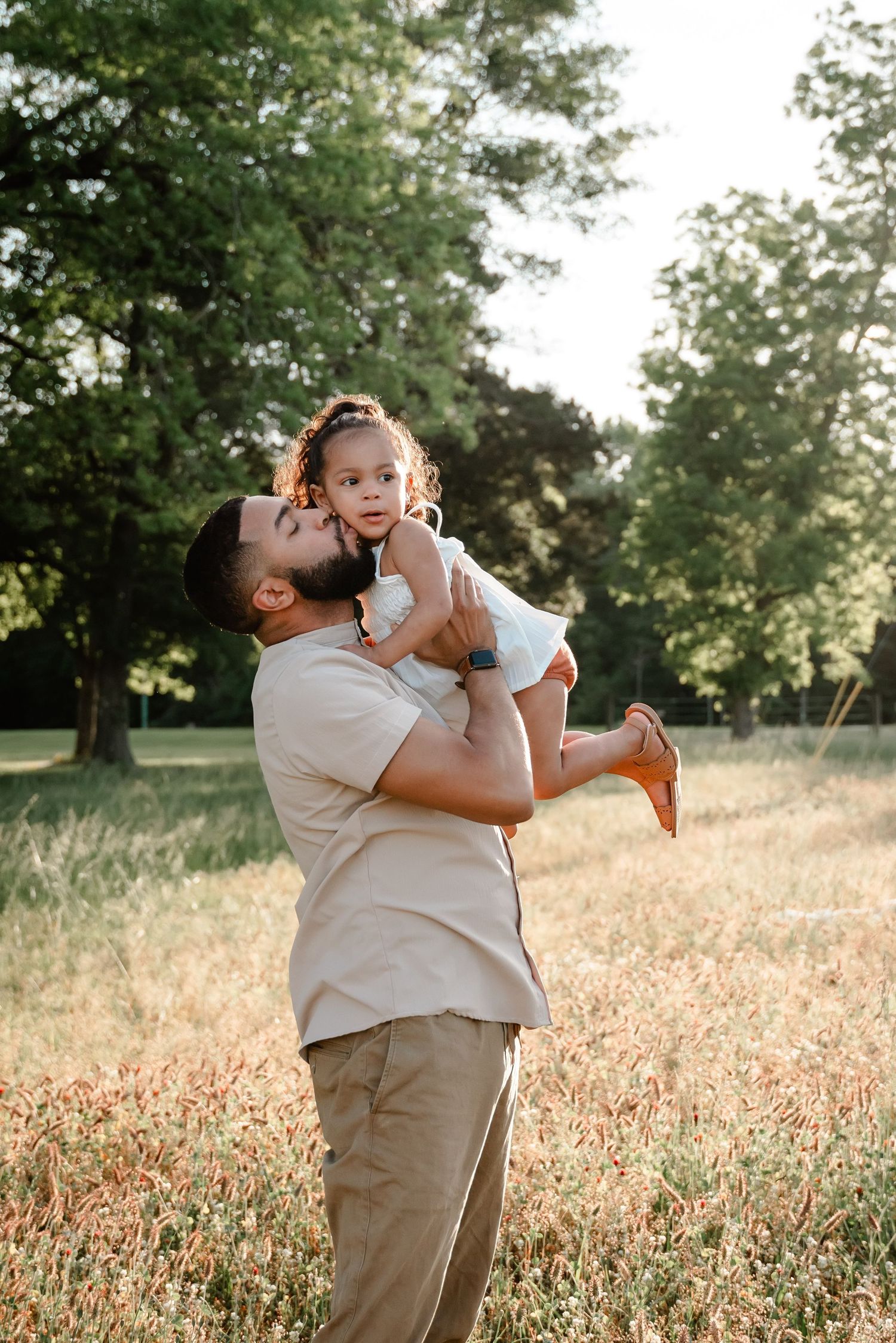 A father lovingly kisses his toddler daughter while lifting her up in a sunlit park meadow.