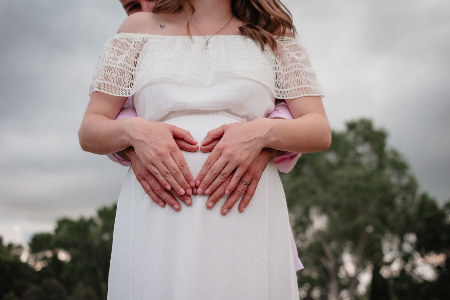 Hands form a heart shape over a pregnant belly in a white lace dress against a cloudy sky and trees.