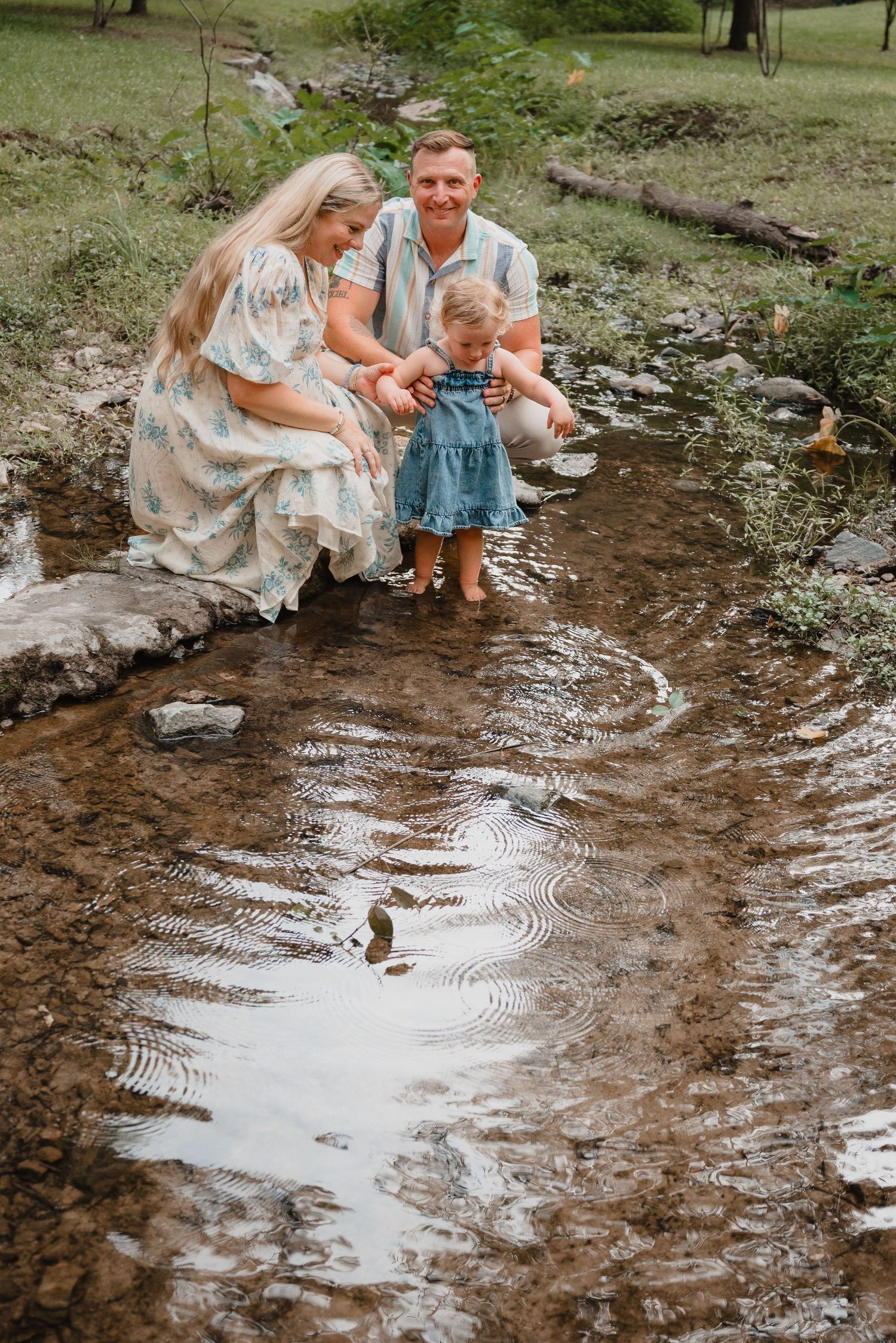 A family of three plays in a shallow creek, with parents helping their toddler splash in the water outdoors.