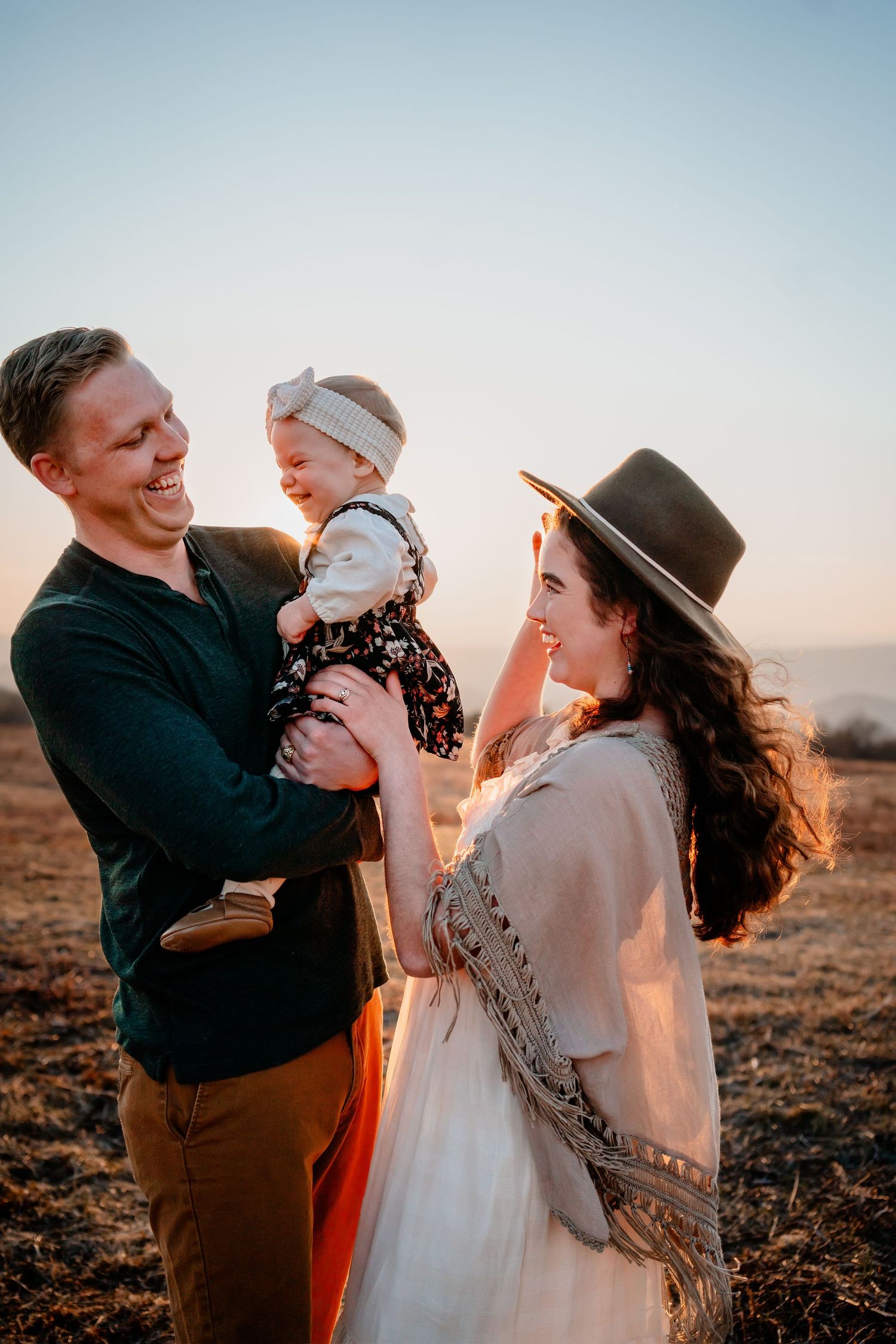 A joyful family of three shares a warm moment at sunset in an open field, with parents smiling at their baby girl.