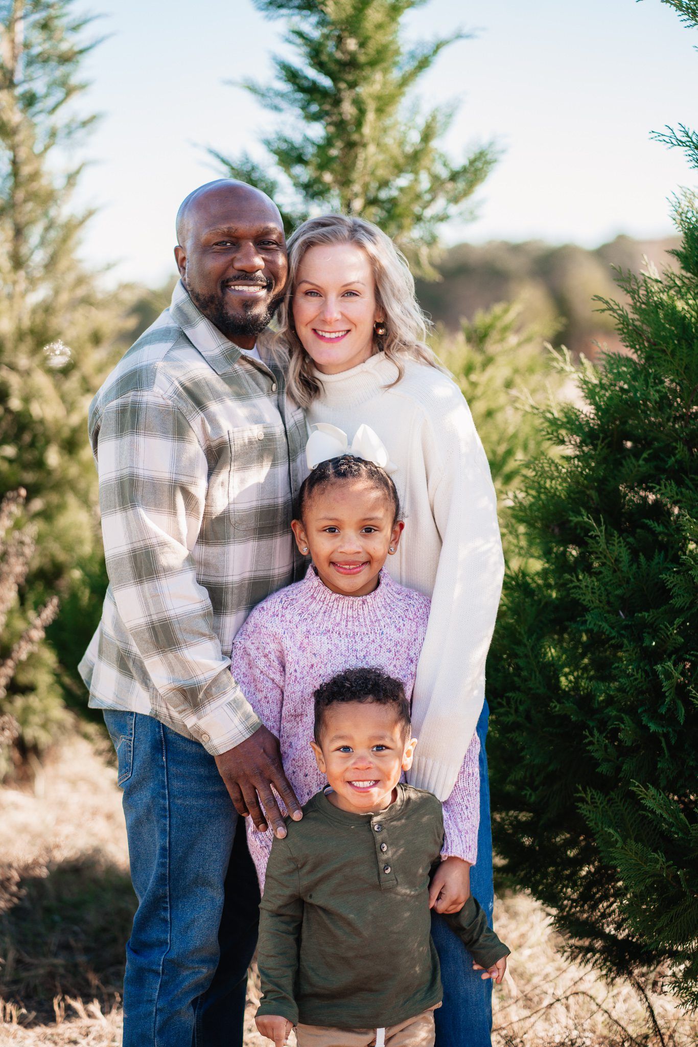 A happy interracial family of four poses among evergreen trees at a Christmas tree farm on a sunny day.