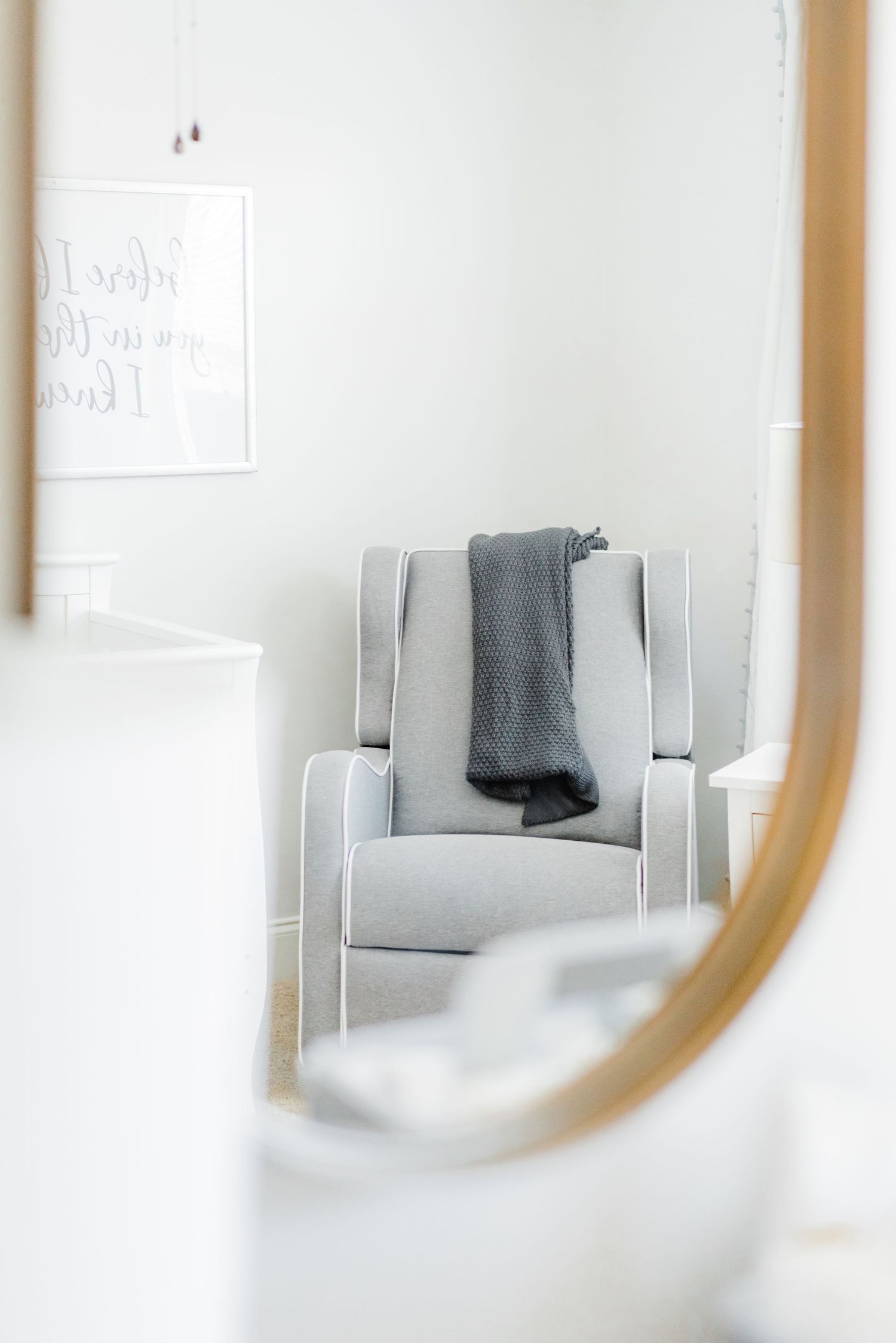 Minimalist nursery interior with round mirror reflecting a gray armchair and white walls.