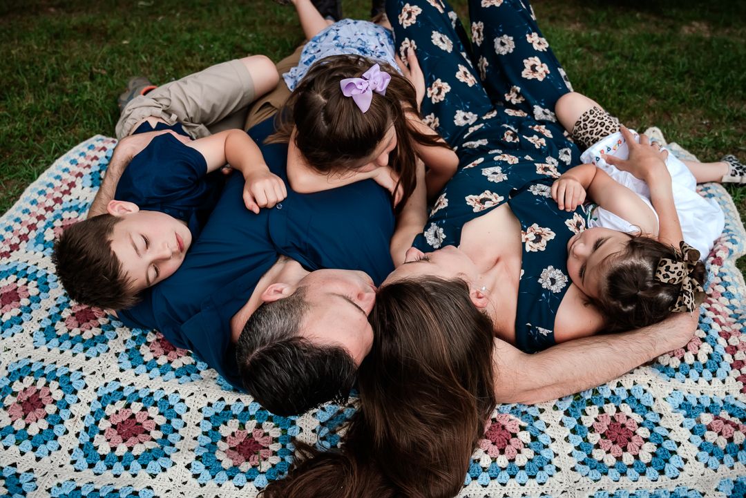 Family members relaxing together on a patterned blanket in the grass during an outdoor photoshoot.