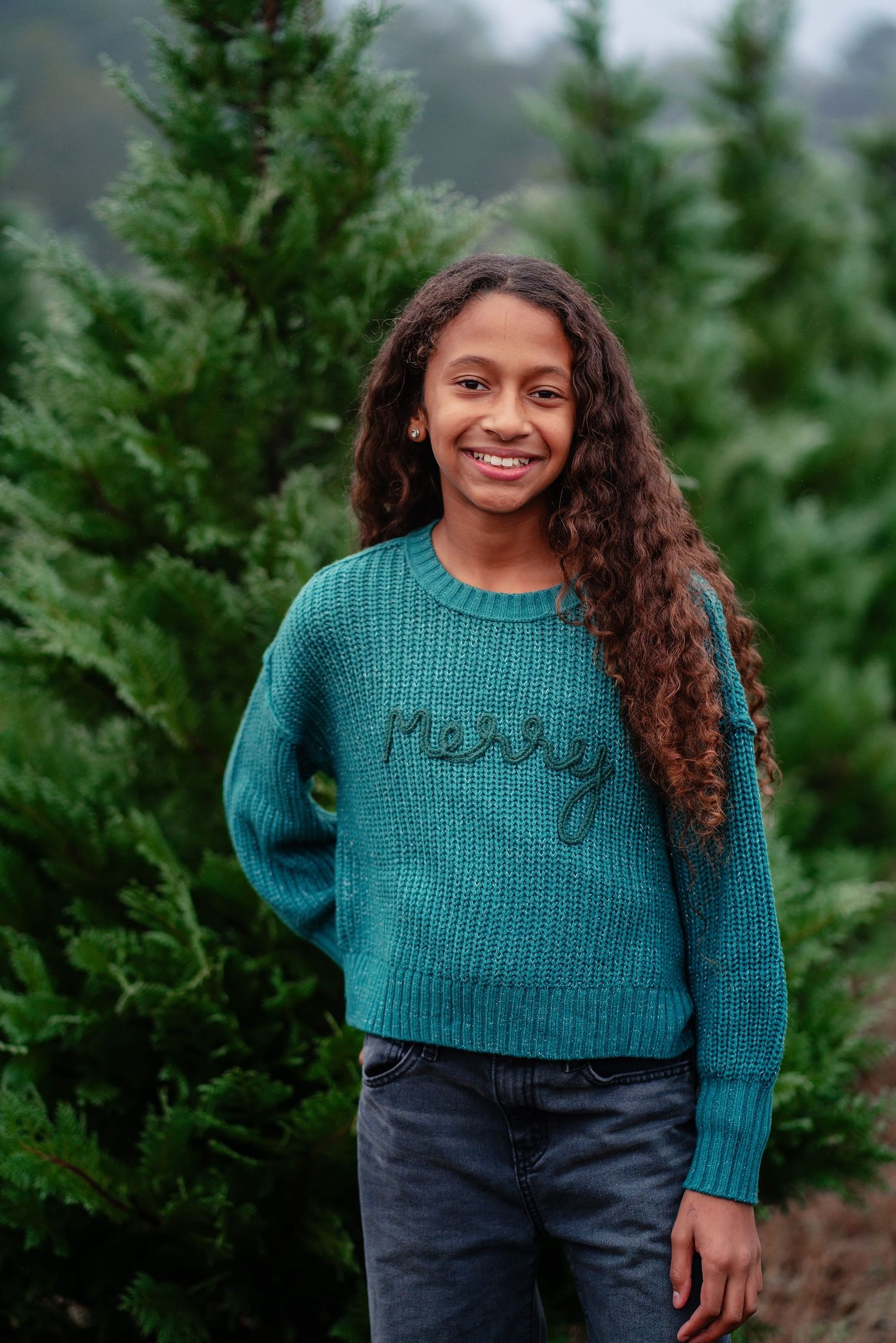 A young person wearing a teal knit sweater stands smiling in front of evergreen trees.
