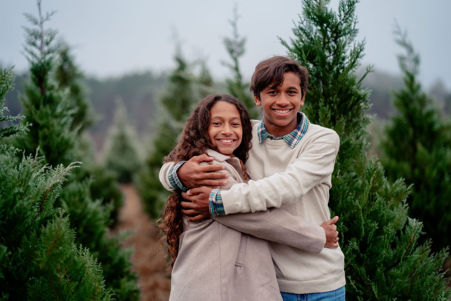 Happy couple embraces while standing among evergreen trees at a Christmas tree farm on a cloudy day.
