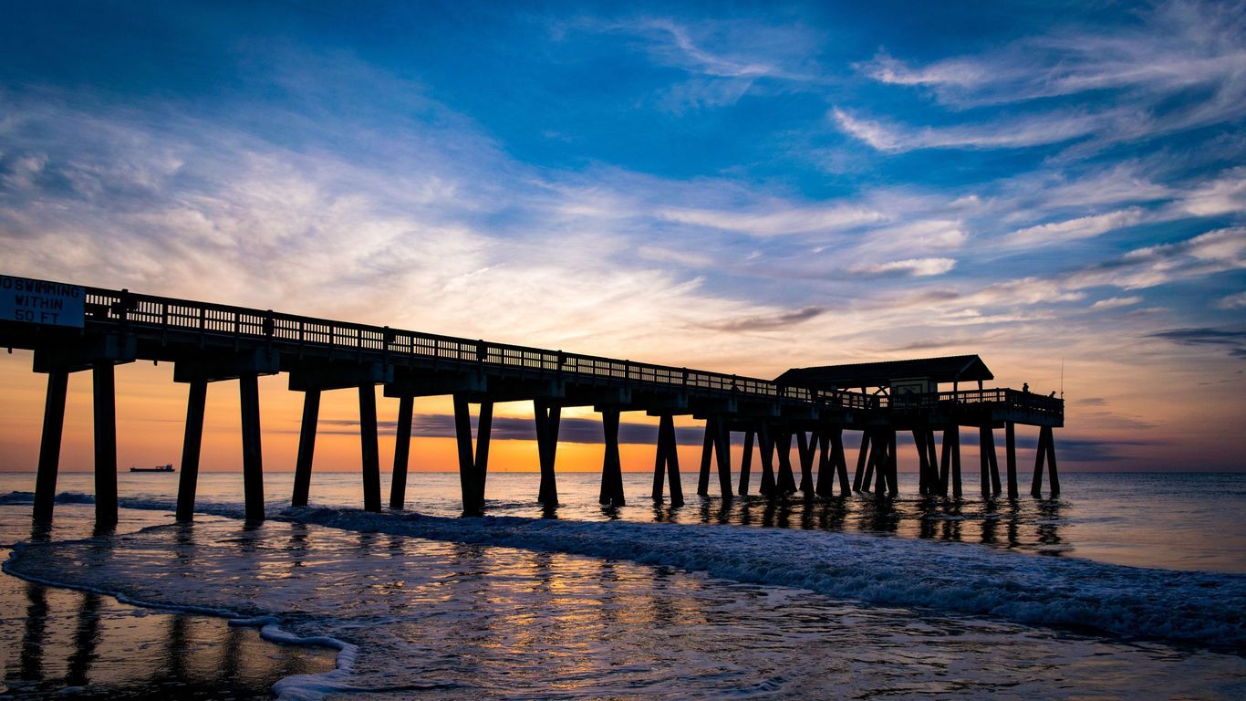 Silhouette of a wooden beach pier at sunset with vibrant blue and orange sky reflecting on calm ocean waves.