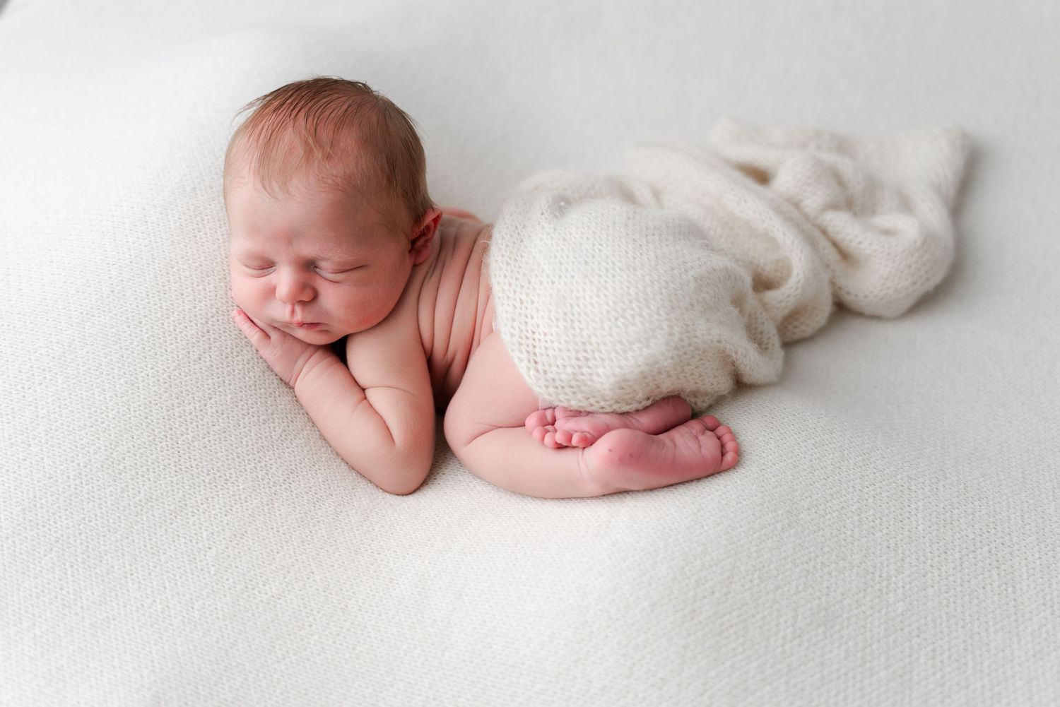 Peaceful newborn baby sleeping posed on white blanket with knit wrap.