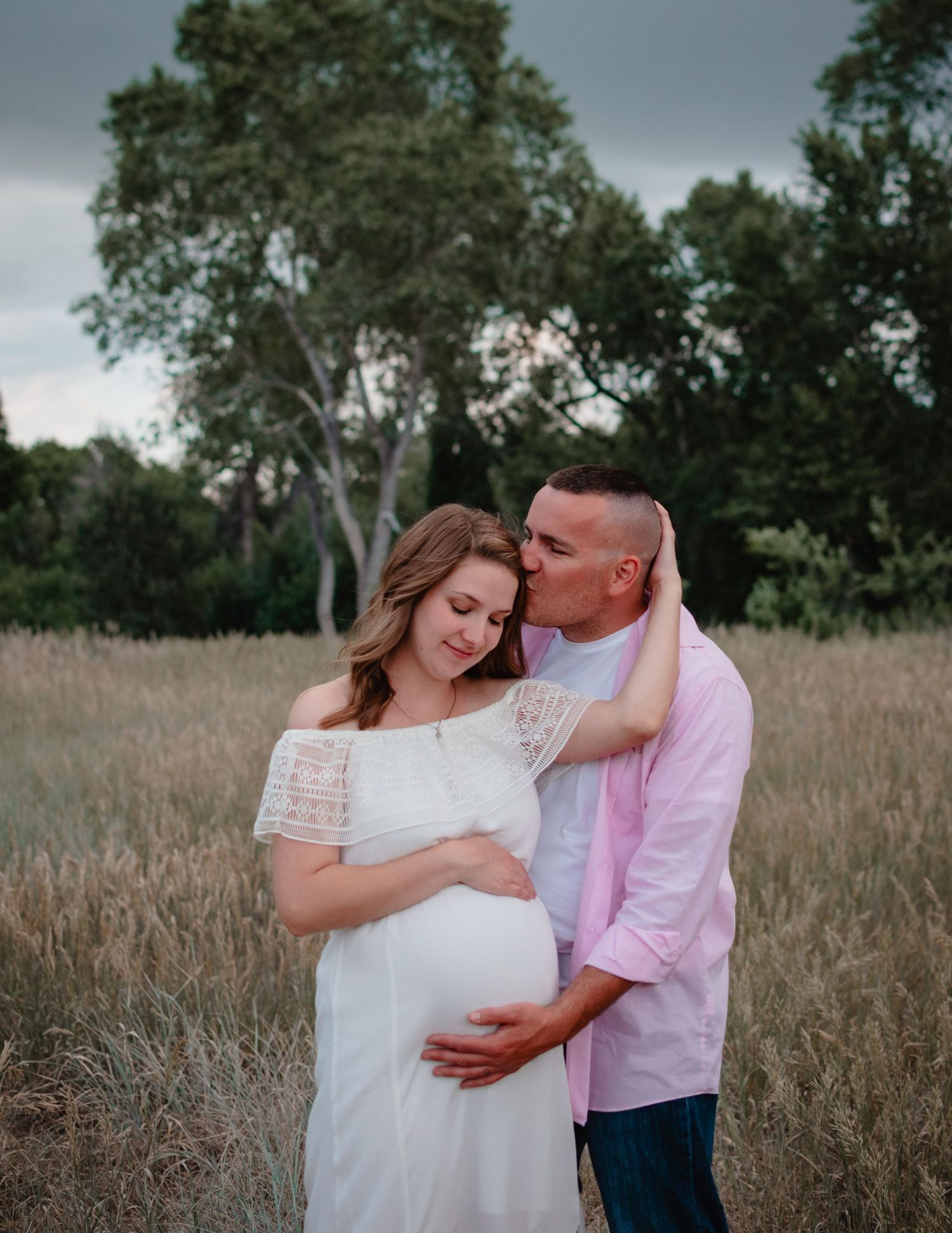 A couple embraces in a rustic field setting during a maternity photo session, wearing white and pink outfits.