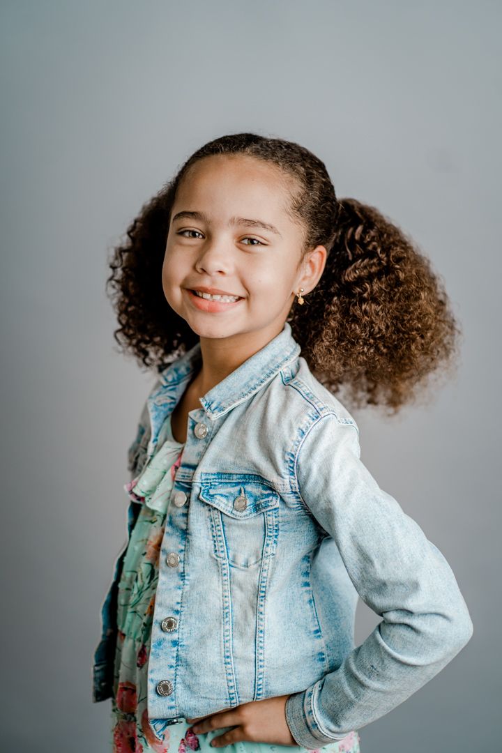 A child in a light denim jacket poses with curly hair against a gray background.