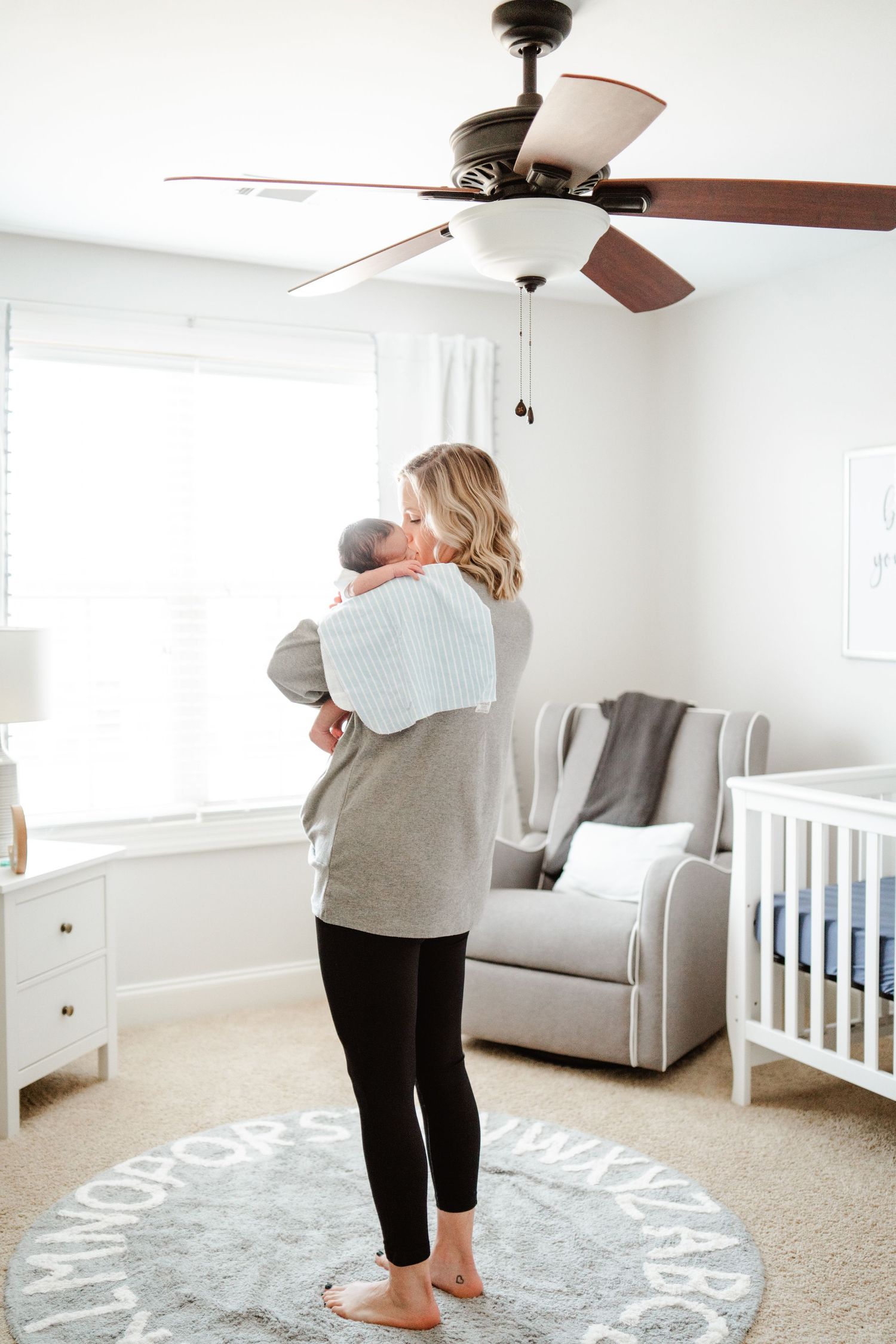 Parent standing in nursery holding baby with ceiling fan and crib visible.
