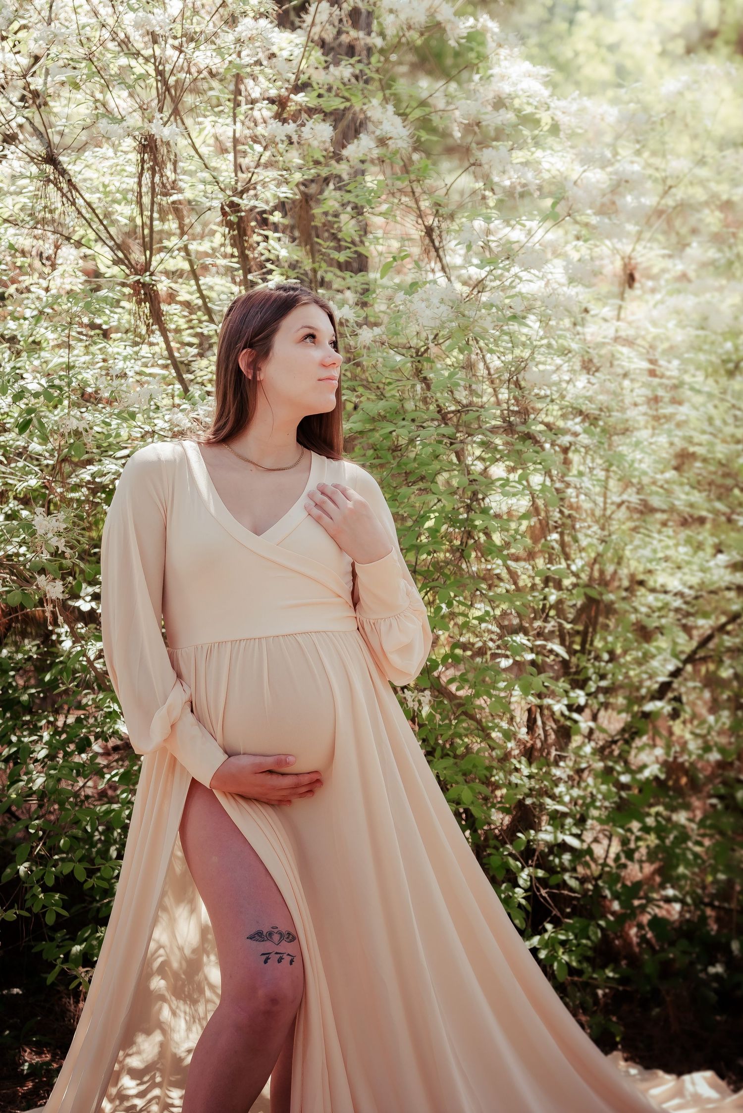 Pregnant woman in elegant beige maternity gown poses outdoors among lush green trees in a serene forest setting.