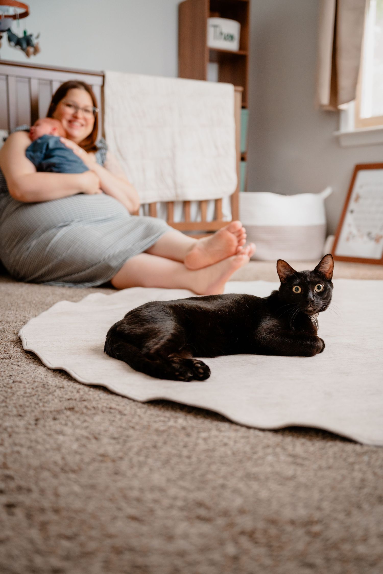 A woman sits on the floor holding a newborn baby in a cozy nursery with cat during lifestyle family photography session Kathleen Georgia