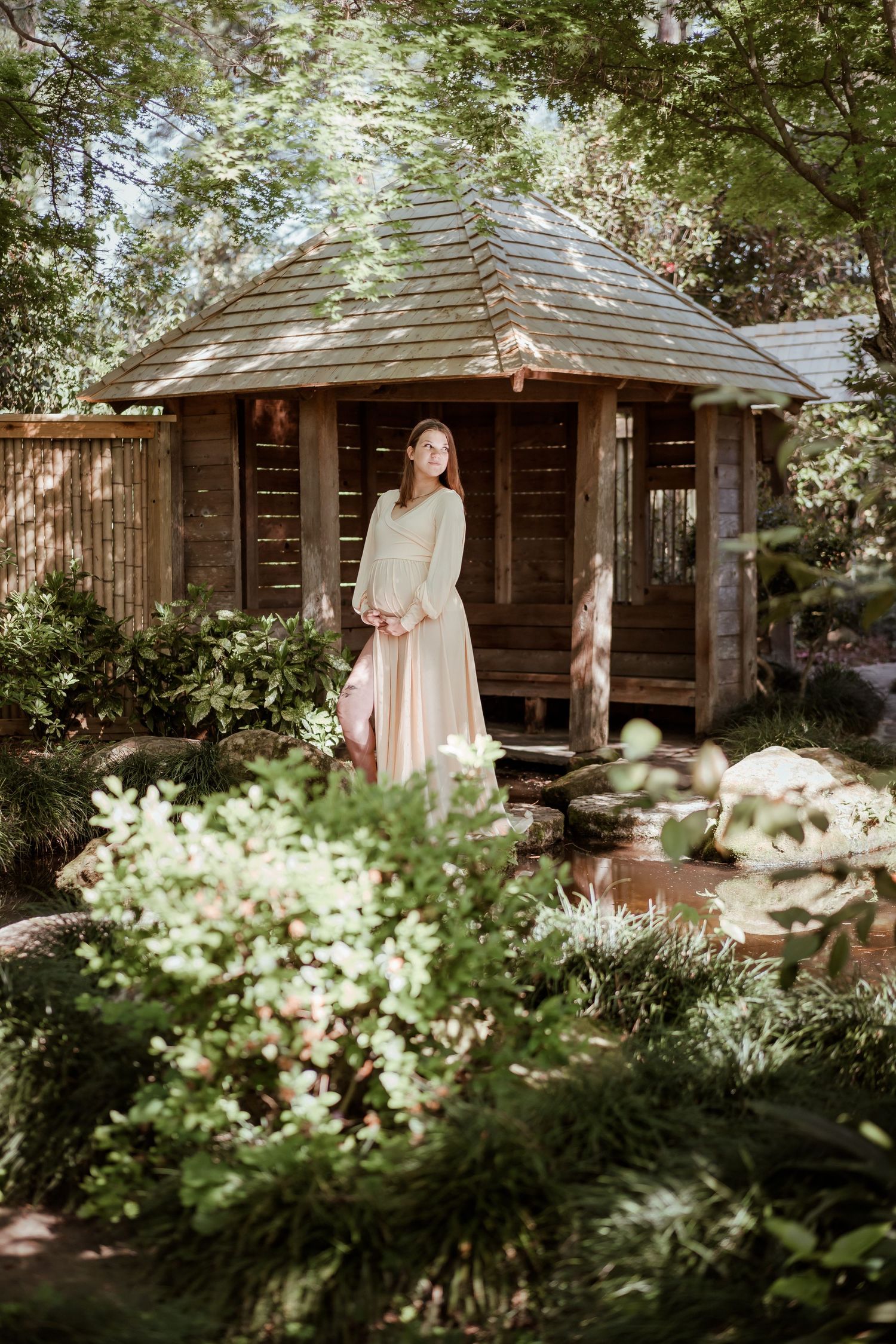 Pregnant woman in flowing pink skirt stands before rustic wooden gazebo surrounded by lush green garden foliage.