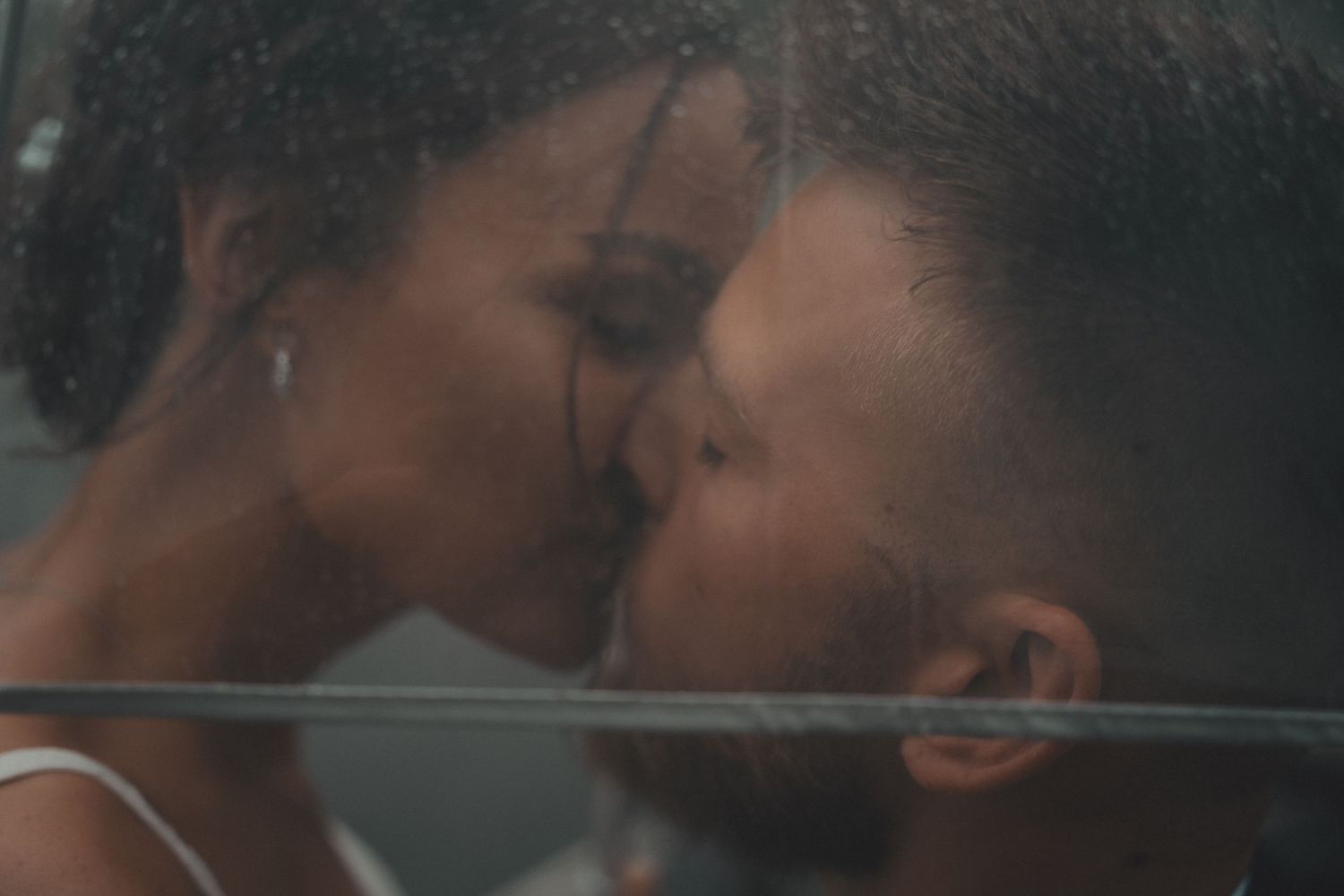 Couple sharing a tender kiss through a rain-covered window in a romantic close-up photo.