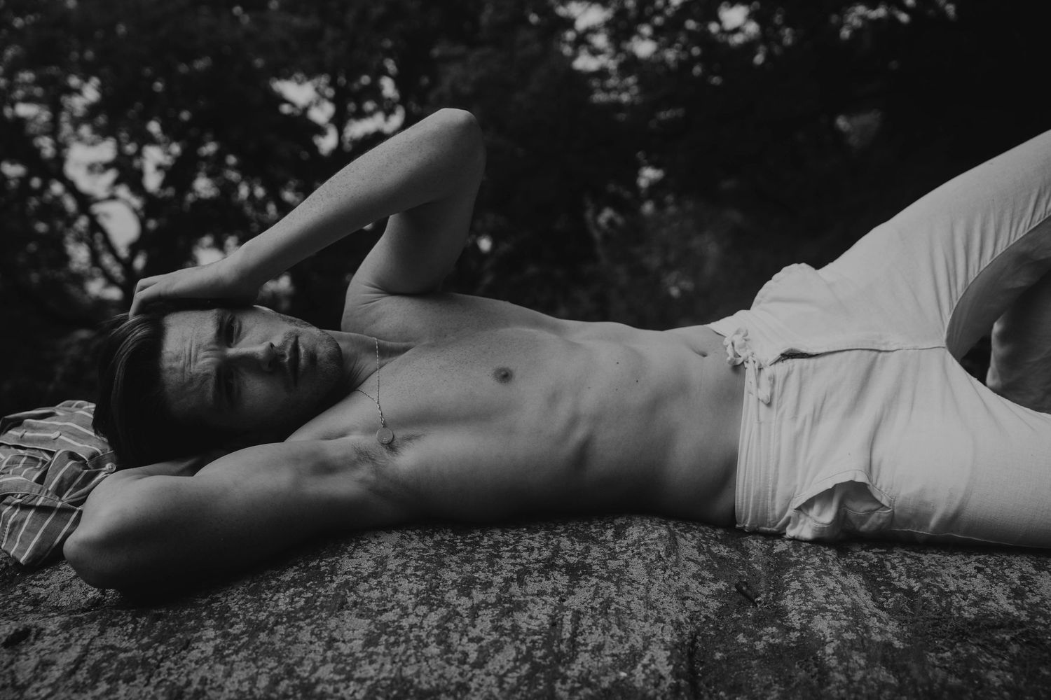 Black and white photo of a shirtless man lying on a rock with one arm raised outdoors.