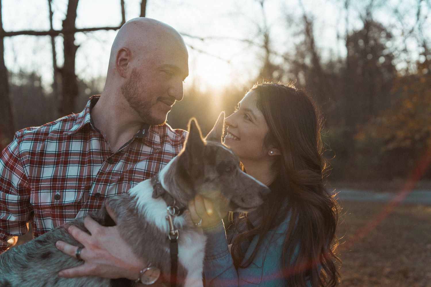 A happy couple shares a romantic moment with their dog at sunset in a wooded setting.