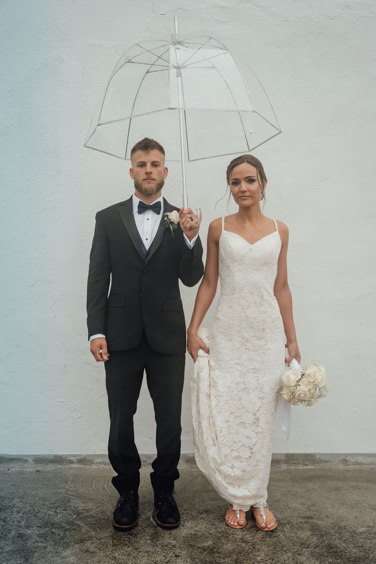 Elegant bride and groom stand under a transparent umbrella against a white wall on wedding day.
