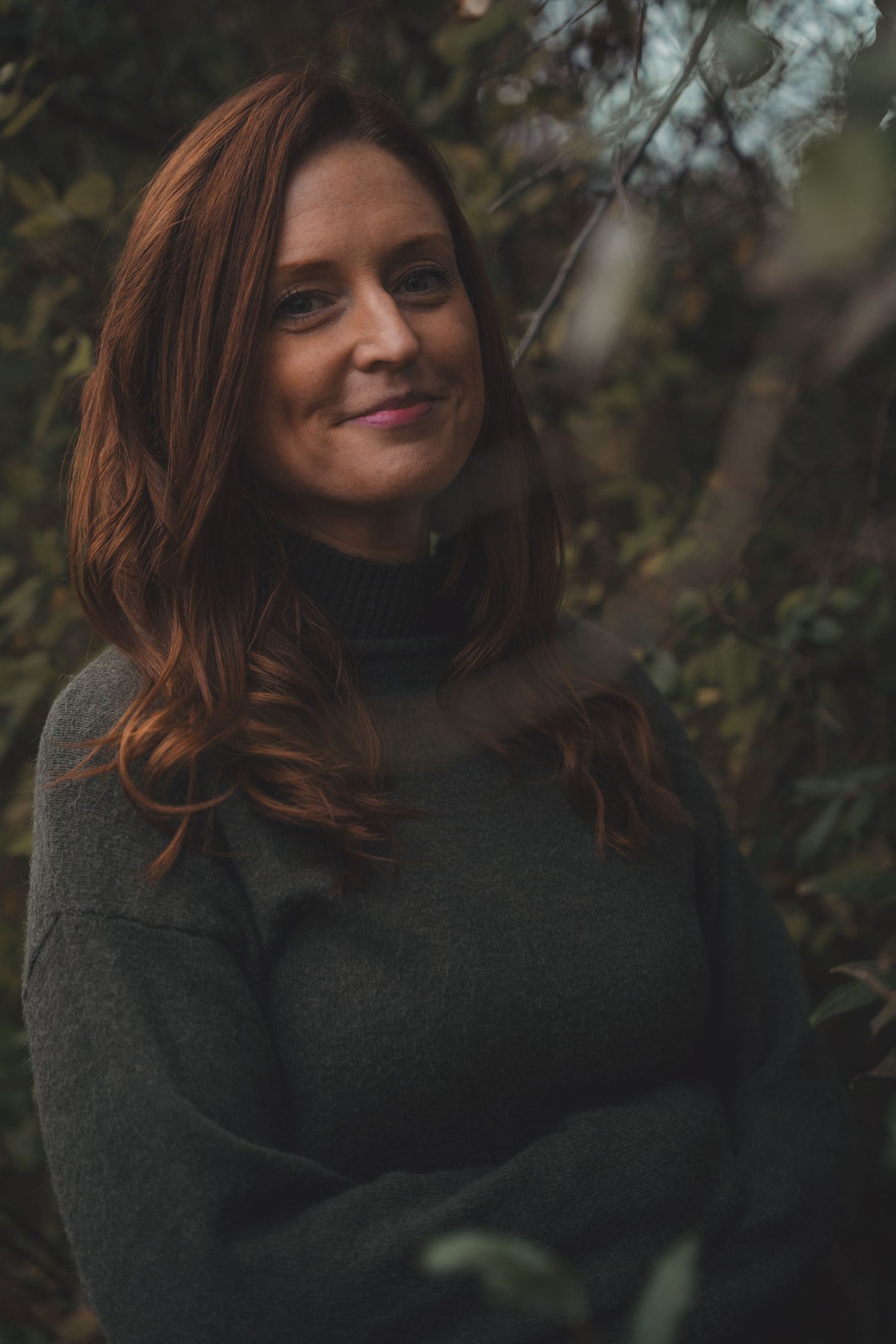 A woman with auburn hair smiles softly in a dark green turtleneck sweater among lush outdoor foliage.
