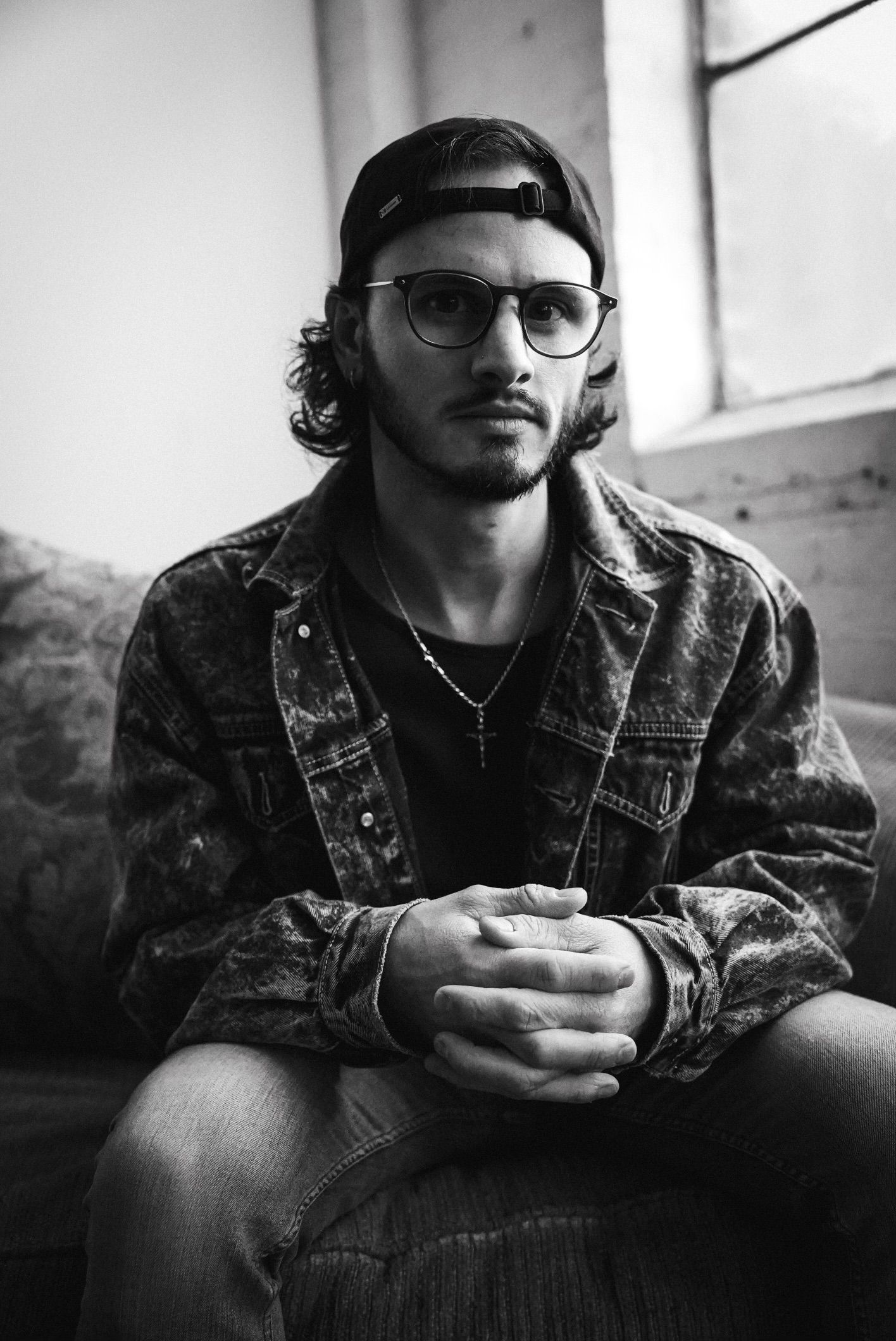 Young man with curly hair, glasses, and backward cap sits with hands clasped in black and white portrait.