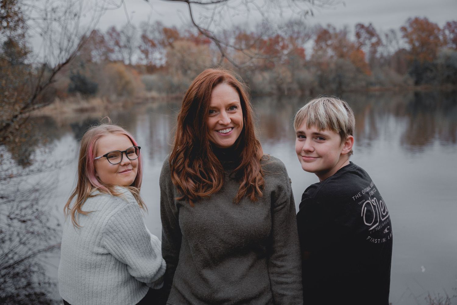 Smiling redhead mother posing outdoors with two children near a lake during autumn.